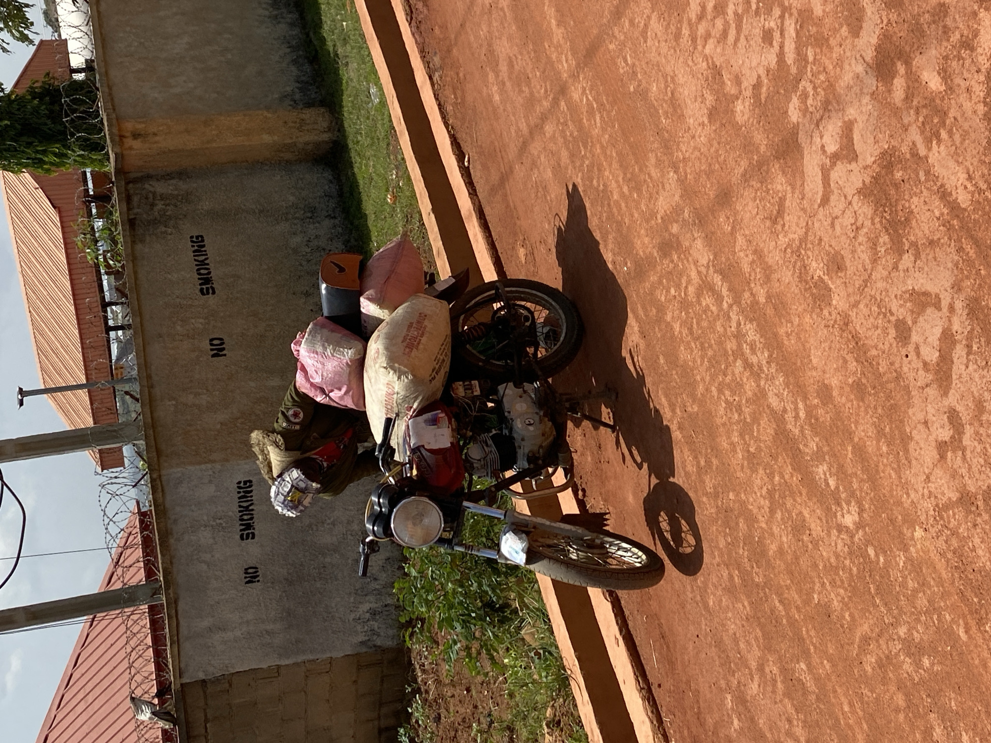 A motorcycle loaded with bags is parked next to a concrete wall with "NO SMOKING" stenciled on it. The motorcycle has several large bags strapped to its rear seat and sides. One of the bags appears to be a white sack with some text on it. The motorcycle itself is a dark color with chrome accents, and it has a headlight and handlebars visible. The scene is outdoors, with a reddish-brown ground surface that appears to be dirt or gravel. Green vegetation is growing around the motorcycle and the wall. Behind the wall, there are barbed wire fences and a building with a reddish-orange roof. The lighting suggests it is daytime, likely sunny, casting shadows from the motorcycle and other objects. The overall impression is of a rural or semi-rural setting in Barakin Angundi, Nigeria, possibly a place where goods are transported by motorcycle. There is no visible human activity or emotion.