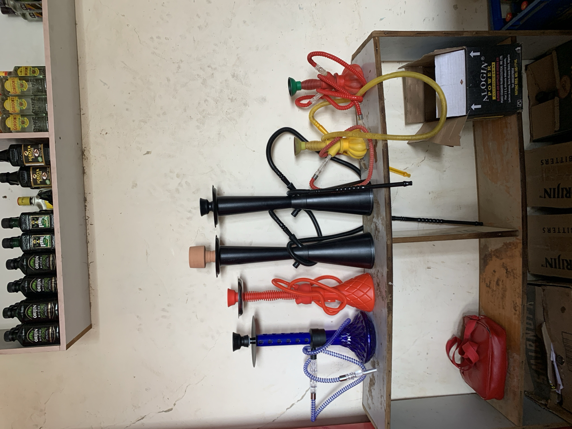 The image shows a shelf in what appears to be a store or storage room, possibly located in the New Stadium area of Nigeria, based on the ALOGIN Power box that is in the image. The shelf contains an assortment of items.
On a pink-edged shelf to the left, multiple bottles of "Gordon's," "Igboya," "Orijin Bitters," and "Federal" are arranged.
Adjacent to this is a wall with a textured surface.
On a set of shelves on the right, there are five hookahs lined up against the wall. From top to bottom, they are: red and green, yellow, two black hookahs, an orange, and a blue. In the back of the shelving unit are multiple boxes labeled “RIJIN BITTERS”, a box labeled “ALOGIN Power Alcoholic Drink”, and a red bag.
