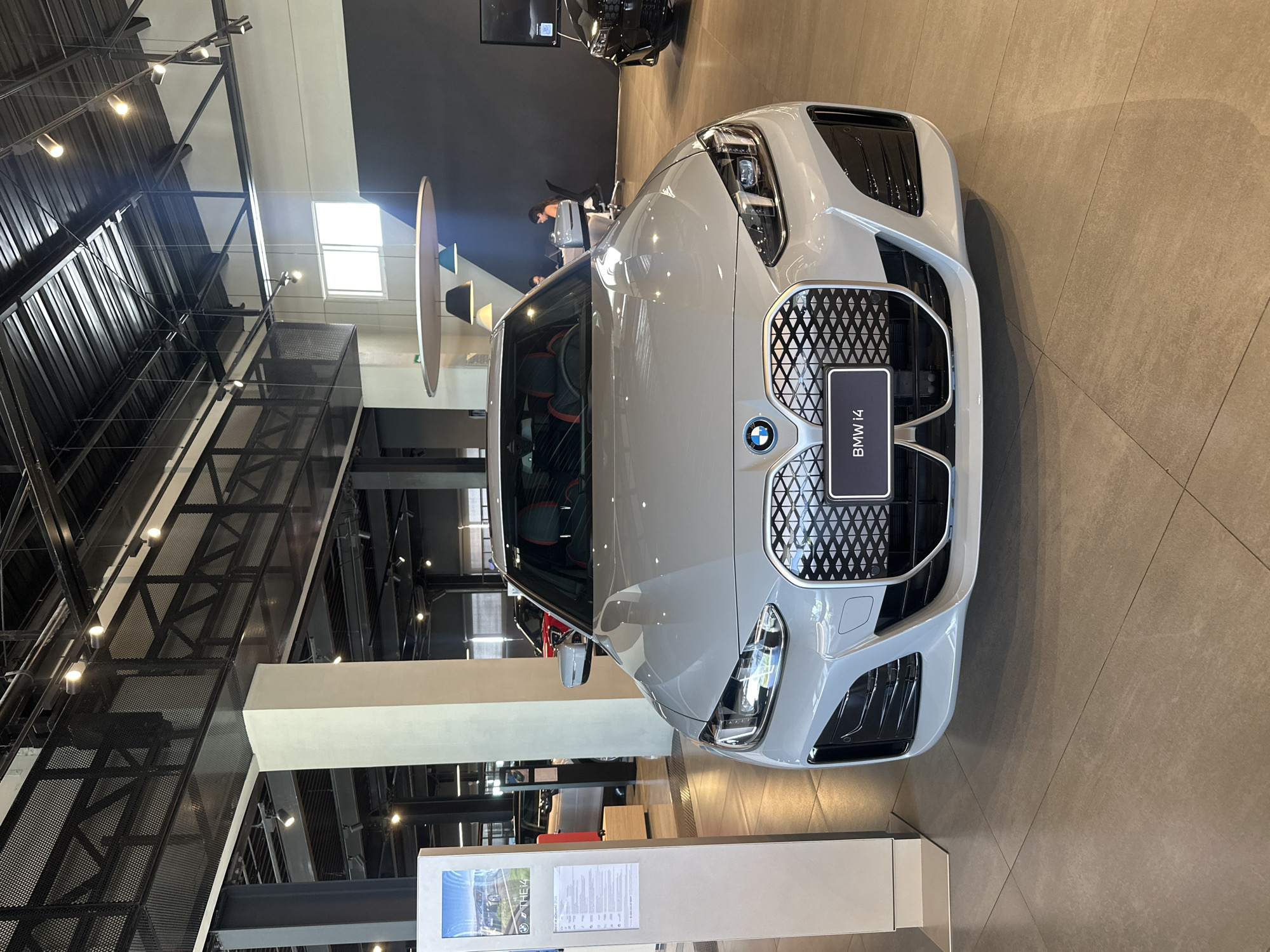 This image captures the interior of a modern car dealership, likely in San Bernardino Tlaxcalancingo, Mexico, showcasing a white BMW i4 sedan. The car is the central focus, positioned in the foreground with its distinctive kidney grille and illuminated headlights prominent. Behind the BMW, other vehicles are visible, including a red car and a black SUV, suggesting a diverse inventory.
The dealership's environment is characterized by sleek, contemporary design. Exposed metal beams and a corrugated metal ceiling create an industrial yet refined aesthetic, illuminated by numerous track lights. A large white pillar stands to the left of the car, and a display stand with information about the BMW i4 is placed next to it. Further in the background, two women are seated at desks, presumably attending to administrative tasks or customer service. The flooring consists of large, light-colored tiles, reflecting the ambient lighting. The overall impression is one of a clean, well-lit, and upscale automotive retail space.