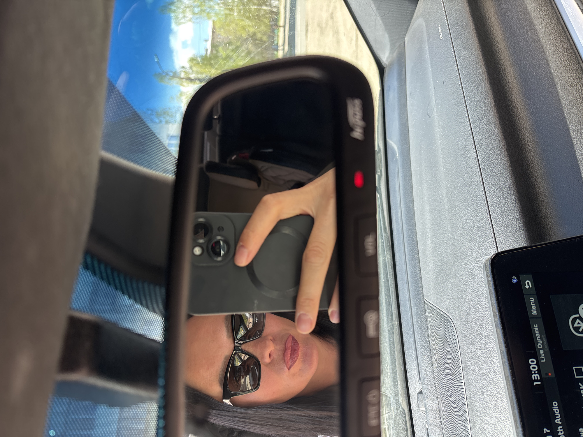 The image shows a person reflected in a car's rearview mirror, holding a smartphone to take a selfie. The person is wearing sunglasses and has dark hair. The car's dashboard and infotainment screen are visible to the right of the mirror. The time displayed on the screen is 13:00. Through the windshield and mirror, a glimpse of the outside environment can be seen. It is impossible to pinpoint the exact location as Chaikovsky, Russia without more details about the environment.