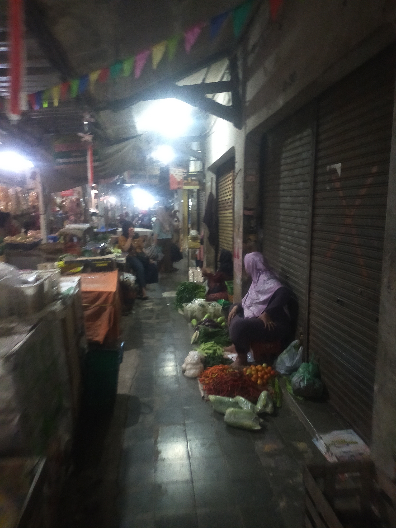 The image depicts a dimly lit market scene. In the foreground, a person wearing a purple hijab and dark clothing is crouched beside a pile of vegetables, including what appear to be green peppers and possibly chilies. Behind them, several metal roll-up doors suggest shops or stalls. The pathway is paved and reflects the overhead lighting. Further down the market aisle, other blurred figures and stalls are visible, with various goods on display. Colorful triangular flags are strung across the ceiling. The overall atmosphere is that of a bustling, indoor market, possibly in the city of Trayeman, Indonesia, given the context provided.