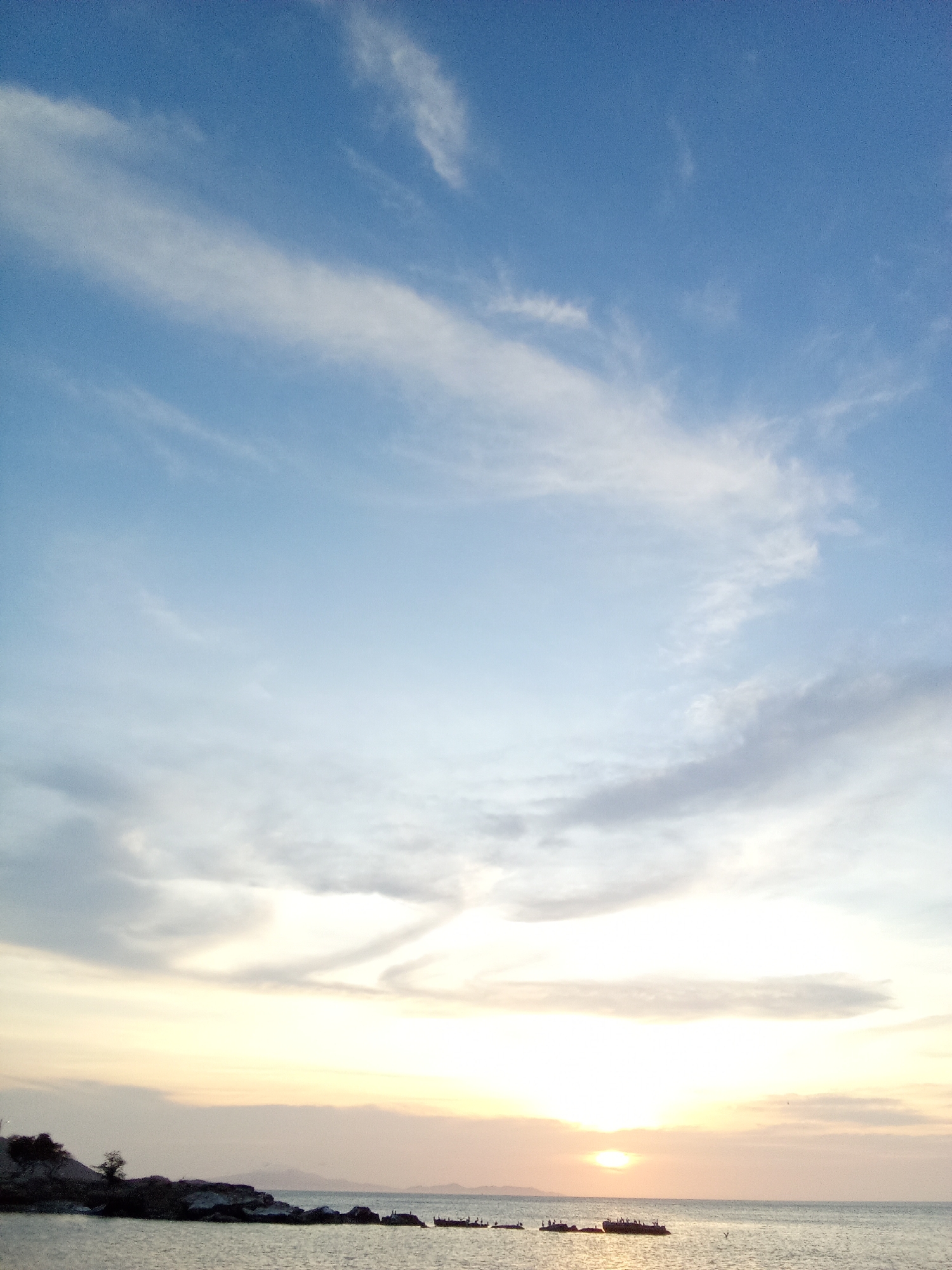 The image shows a sunset over the ocean, likely near Juan Griego, Venezuela. The sky is predominantly blue with wispy white clouds. Closer to the horizon, the sky transitions to hues of yellow and orange. The sun is setting directly on the horizon, appearing as a bright spot. In the foreground, there is a rocky shoreline with some vegetation. Several small boats are visible on the water, some appearing to have people on them. In the distance, there's a faint suggestion of hills or mountains.