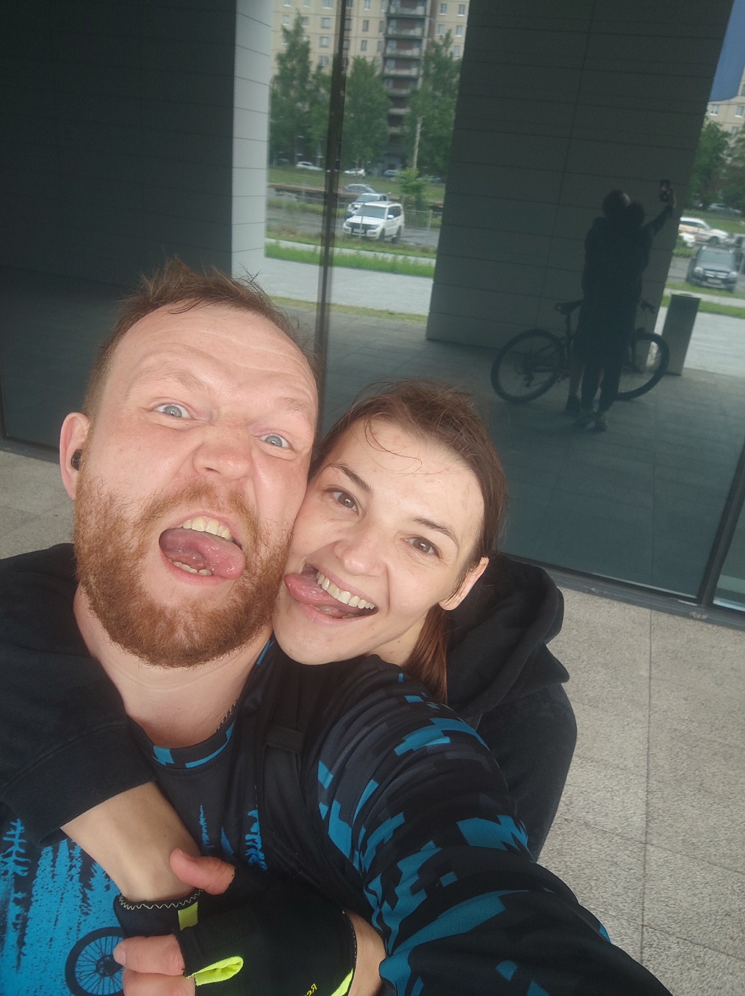 A man and a woman are taking a selfie in front of a building with reflective glass windows. The man, who is on the left, has a beard and is sticking his tongue out. The woman, to his right, is also sticking her tongue out. In the reflection of the glass, a person is visible holding a phone, taking a picture of themselves with a bicycle behind them. The background shows a street with cars and buildings. The scene appears to be in an urban environment, possibly in Saint Petersburg, Russia, given the architectural style of the building and the presence of urban infrastructure. The people are dressed in athletic wear. The man has on a black hoodie with blue accents and the woman is wearing a black hoodie. The man is wearing a black glove on his right hand.
