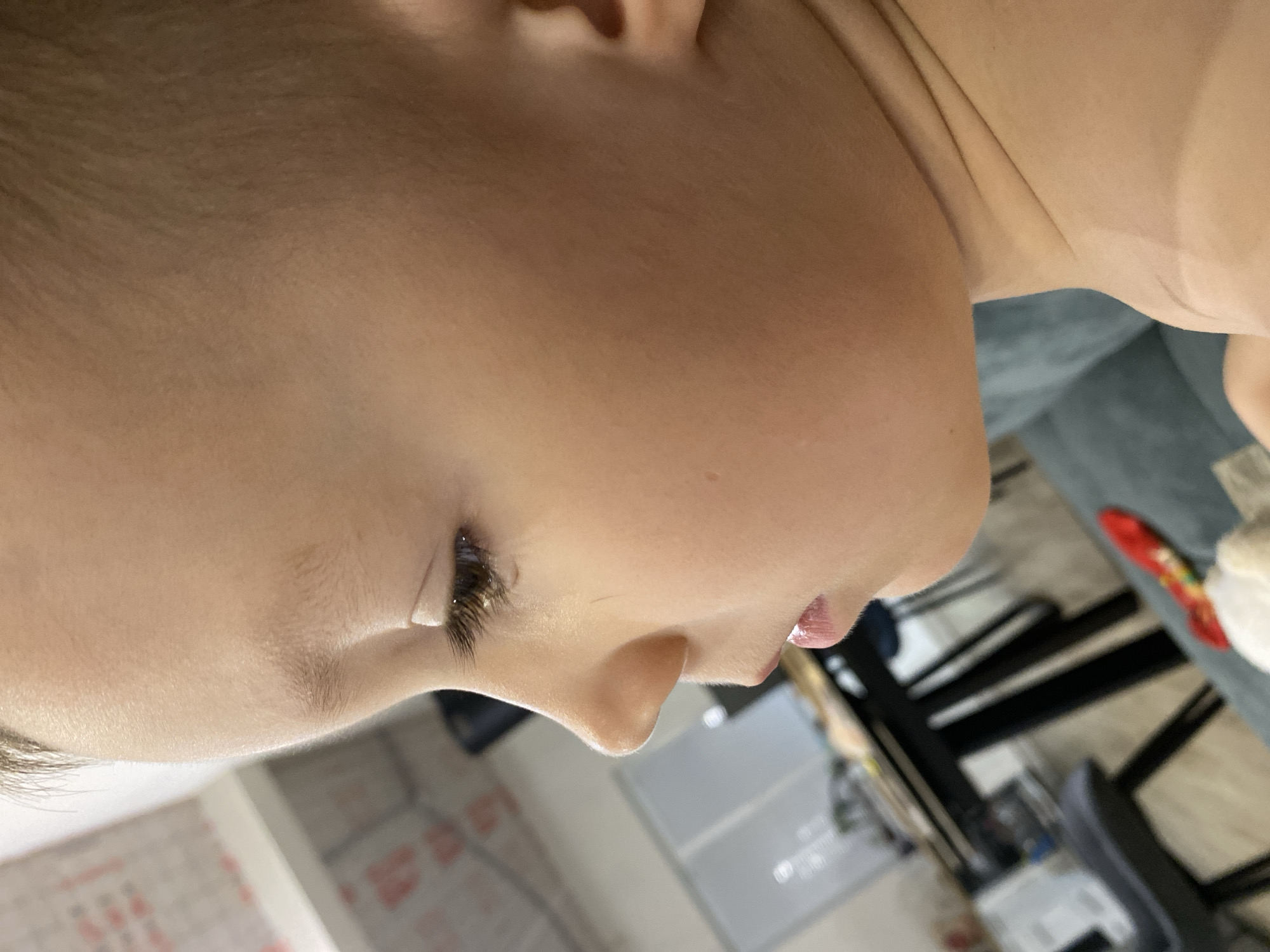 A close-up, vertical shot captures the left profile of a young person, likely a child. The subject's light-toned skin, ear, and the side of their head are prominent, with light-colored hair visible at the top and front. Their eye is partially visible, featuring long, dark eyelashes that appear to have a slight gloss or sheen. Below the eye, a distinct nose bridge leads to a small, slightly upturned nose. The lips are visible, appearing full and slightly parted, also with a glossy texture. The person is wearing a light-colored top or clothing that blends with their skin tone.
The background is out of focus but indicates an indoor setting. On the right side, a blue or grey sofa is discernible, with a red object, possibly a toy or snack wrapper, resting on it. Further back, indistinct dark-framed furniture, possibly chairs or a table, and blurred light-colored surfaces suggest a room, likely a living area or dining space. There is no clear activity depicted, and the person appears calm. No text is visible within the image.
