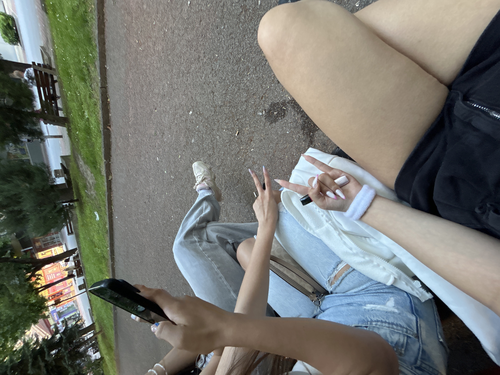 The image depicts a close-up view of several individuals, focusing on their lower bodies and hands, seated on a park bench. The environment suggests an urban park setting with a paved path, grass, trees, and buildings visible in the background. The individuals are dressed casually in jeans and athletic wear. One person is holding a smartphone, while another is making a peace sign with their fingers and wearing a white wristband. Another person's arm with long, manicured nails is also visible. The overall scene conveys a relaxed, social gathering in a public outdoor space, potentially in the city of Shumen, Bulgaria, given the context provided.