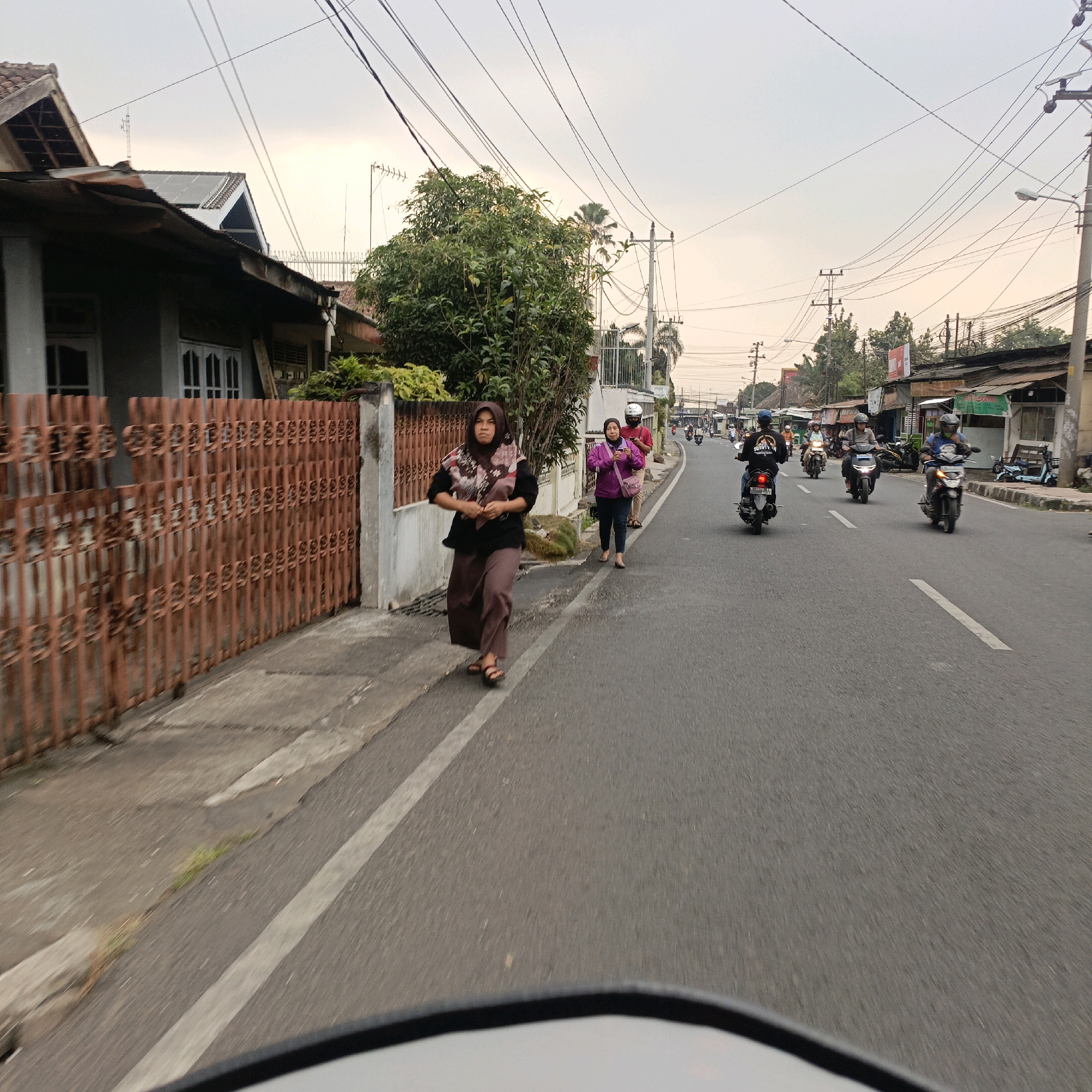 This image depicts a street scene in Salatiga, Indonesia, likely taken from a moving vehicle. In the foreground, the edge of a vehicle's windshield or mirror is visible. The road is asphalt with a white dashed line marking the lane. To the left, a person wearing a hijab and a brown and black patterned scarf, along with a long brown skirt and sandals, walks along the sidewalk. They are holding something in their hands. Behind them, another person in a purple jacket and dark pants walks on the sidewalk. On the road, several motorcycles are visible, some with riders, traveling in the same direction as the viewer. One motorcycle in the middle ground has a rider wearing a black helmet and a black shirt with white text that appears to say "SUFISM." To the left of the sidewalk, there is a reddish-brown metal fence in front of a building with a tiled roof. Trees and power lines are visible in the background, along with more buildings and shops on the right side of the street. The sky is overcast with a pale yellow hue, suggesting either early morning or late afternoon.