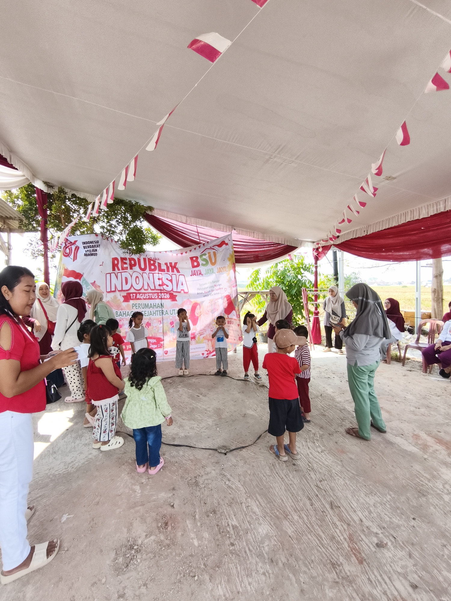 This is a full-shot, eye-level, outdoor photograph taken under a white tent decorated with red and white Indonesian flags. In the center of the image, a group of children and adults are gathered around a circular rope drawn on the ground. Some children are participating in an activity, with one child covering their eyes. A large banner behind them reads "REPUBLIK INDONESIA" and "17 AGUSTUS 2026". Several adults are observing the children. The ground is uneven dirt and concrete. In the background, there are trees and a field of dry grass. The setting appears to be a community event, likely a celebration of Indonesian Independence Day. The location is the city of Bandar Lampung, Indonesia.