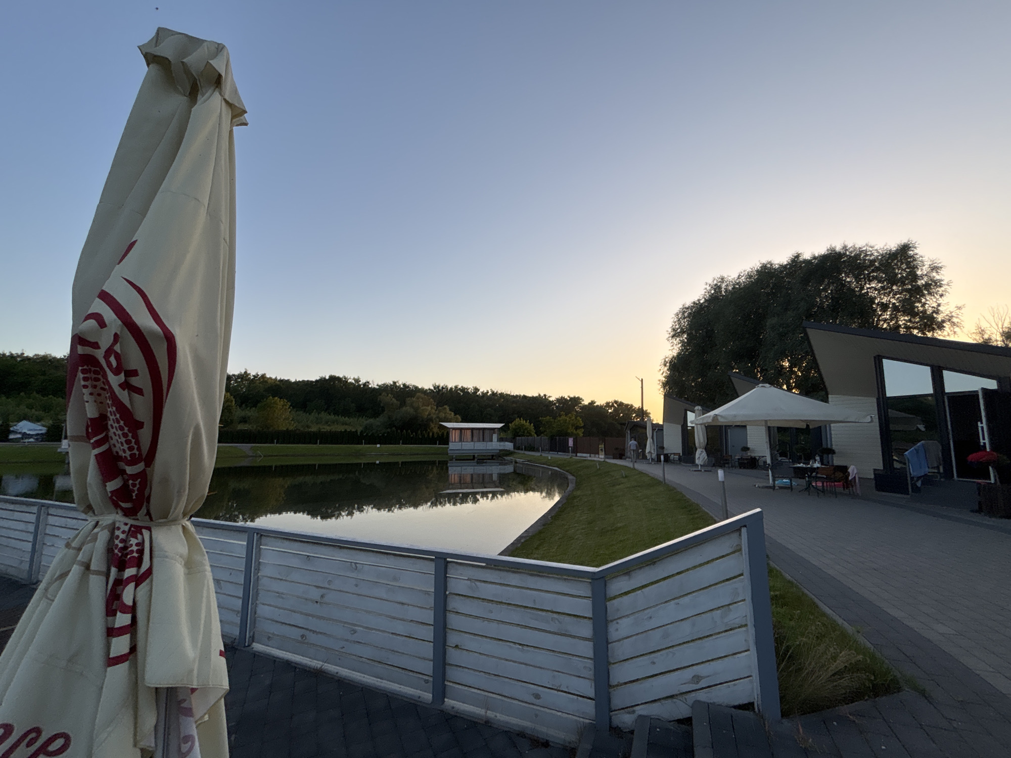 The image shows an outdoor scene at dusk. In the foreground, a large, closed umbrella with a red and white logo stands to the left. A light-colored wooden fence runs along a paved walkway. Beyond the fence, a calm body of water reflects the sky and surrounding trees. On the far side of the water, a small, modern building with large windows sits on stilts, and a line of trees forms the background. To the right of the water, more buildings with outdoor seating areas are visible, along with large umbrellas providing shade. The sky is a clear gradient of pale blue to a soft yellow near the horizon, indicating sunset or sunrise. A few people are subtly visible in the distance, walking along the path and near the buildings. The overall mood is peaceful and serene. No discernible text is clearly readable.