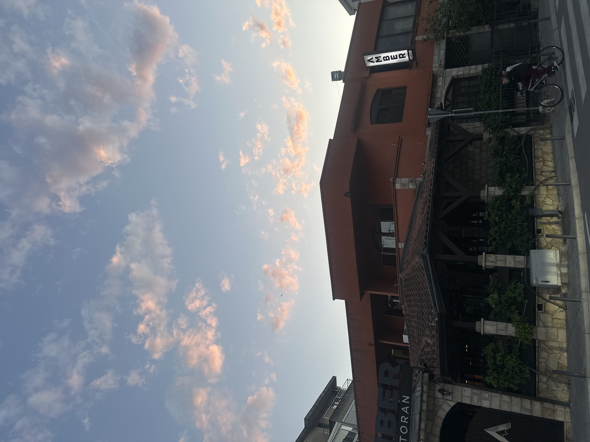The image shows a view of the Amber Restoran in Budva, Montenegro, with a partial view of the sky filled with pink-tinged clouds. The restaurant is housed in a multi-story building with a reddish-brown facade. A sign with the name "AMBER" is visible on the upper part of the building. A cyclist is seen partially obscured on the sidewalk next to the building. The lower portion of the restaurant features a covered patio with tiled roofing and stone detailing. A street runs along the right side of the building.