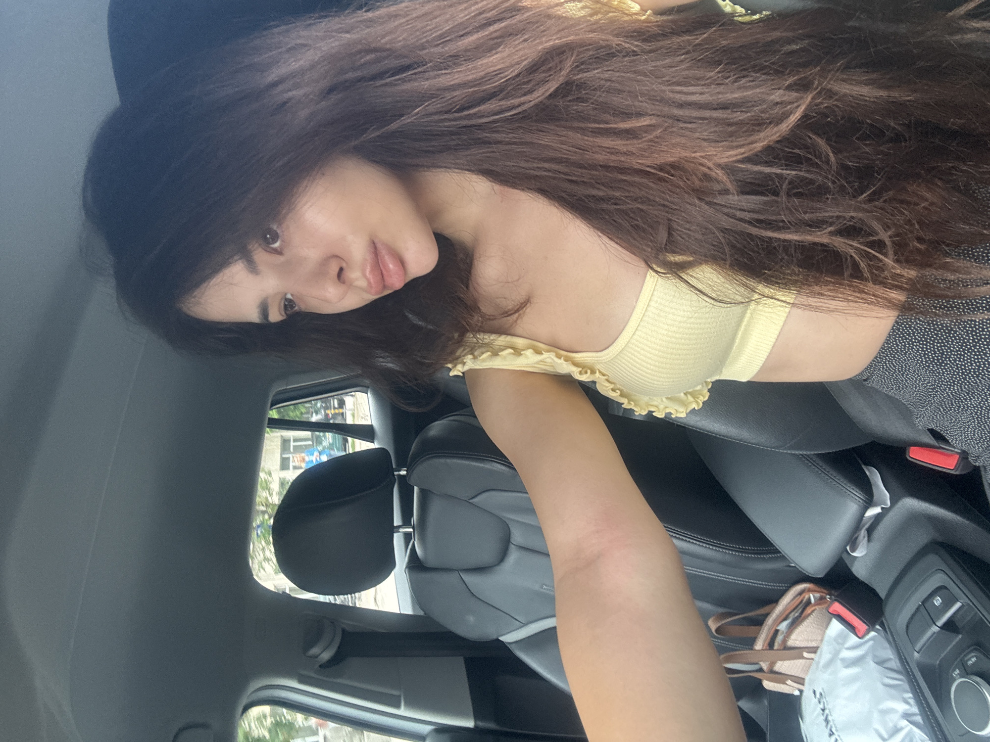 A young woman with long, dark brown, wavy hair is captured in a selfie or portrait taken inside a car. Her head is tilted slightly, and she looks directly into the camera with a neutral to slightly pensive expression, her lips slightly parted. She is wearing a light yellow, ribbed crop top with ruffled details along the neckline, paired with a dark-colored, possibly patterned skirt or bottom.
The setting is the interior of a modern car, likely from the driver's or front passenger's perspective. Black leather seats are visible, along with a center console featuring various controls. Through the car windows, blurred architectural structures and green foliage suggest an urban or suburban environment, consistent with the location context of Plovdiv, Bulgaria. The bright lighting indicates it is daytime.
On the passenger seat or floor, a light brown/beige handbag and a white bag are visible. The white bag has partially discernible text, appearing to read "LANCC" or "LANCE". The overall scene depicts a casual moment of the woman posing inside a stationary vehicle.