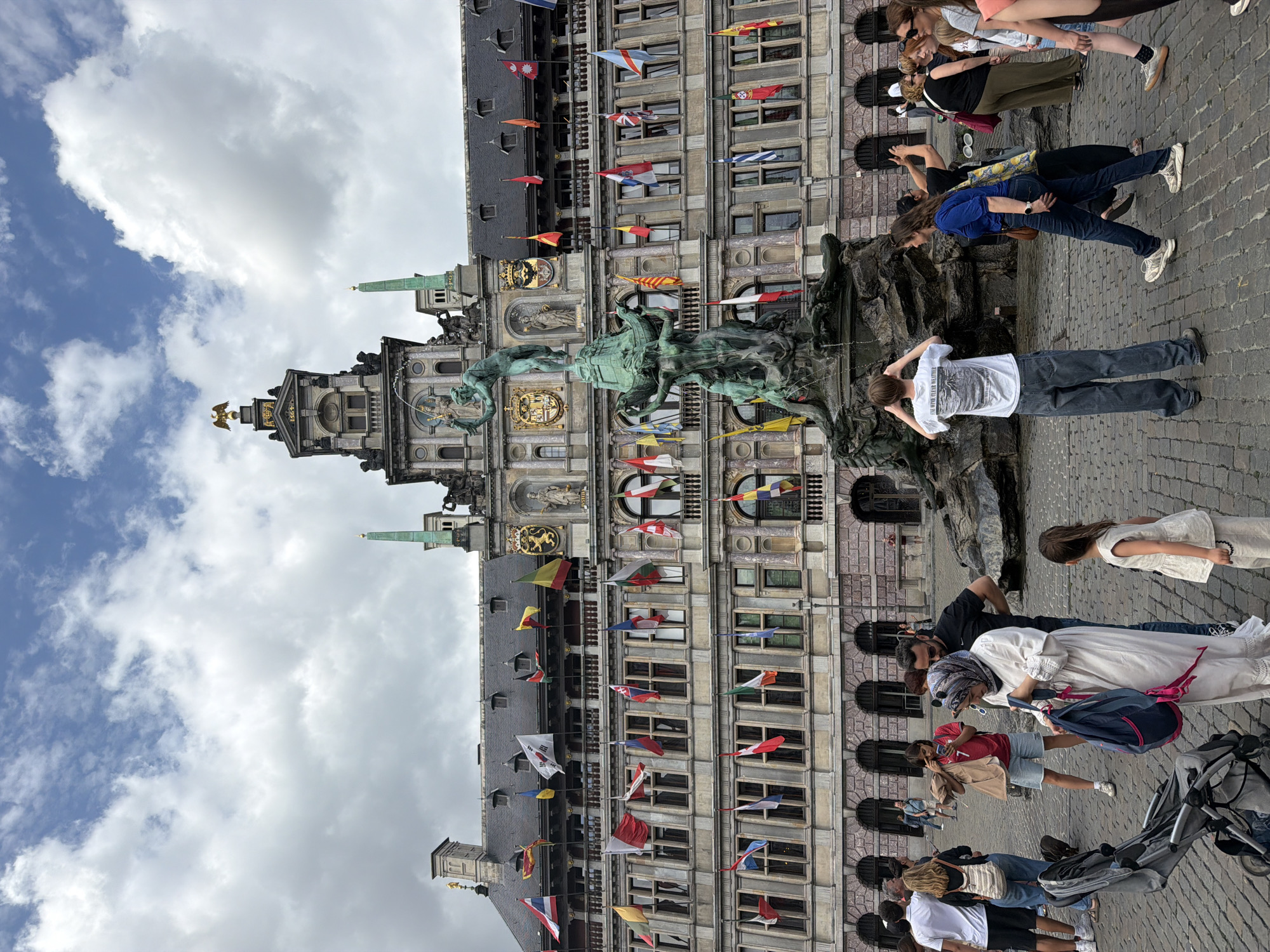 The image depicts a bustling public square in front of the Antwerp City Hall, a prominent Renaissance building adorned with numerous flags of different nations. In the foreground, a large bronze fountain featuring mythological figures is a central element. A diverse group of people are present, engaged in various activities such as walking, taking photos, and observing the surroundings. A baby stroller is visible in the lower left corner. The sky is partly cloudy, suggesting dynamic weather conditions. The cobblestone pavement indicates a historic urban setting. The overall scene captures a lively atmosphere at a landmark location.
