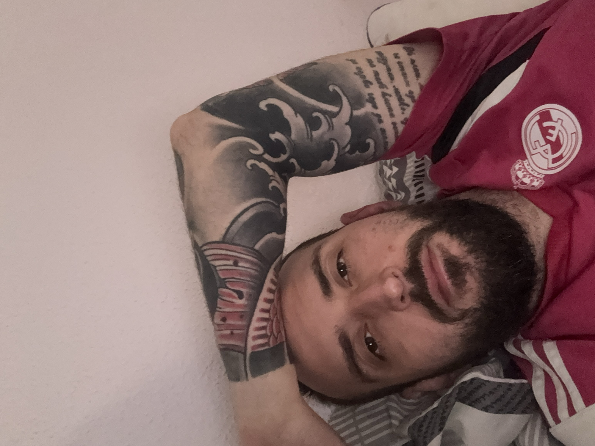 A man is lying down, face up, with his head resting on a pillow. He has dark hair, a beard, and is wearing a red shirt with a white logo on the right chest, and white stripes on the sleeve. He has multiple tattoos on his left arm. The background is a white wall. It is not possible to determine if this image was taken in Soria, Spain.