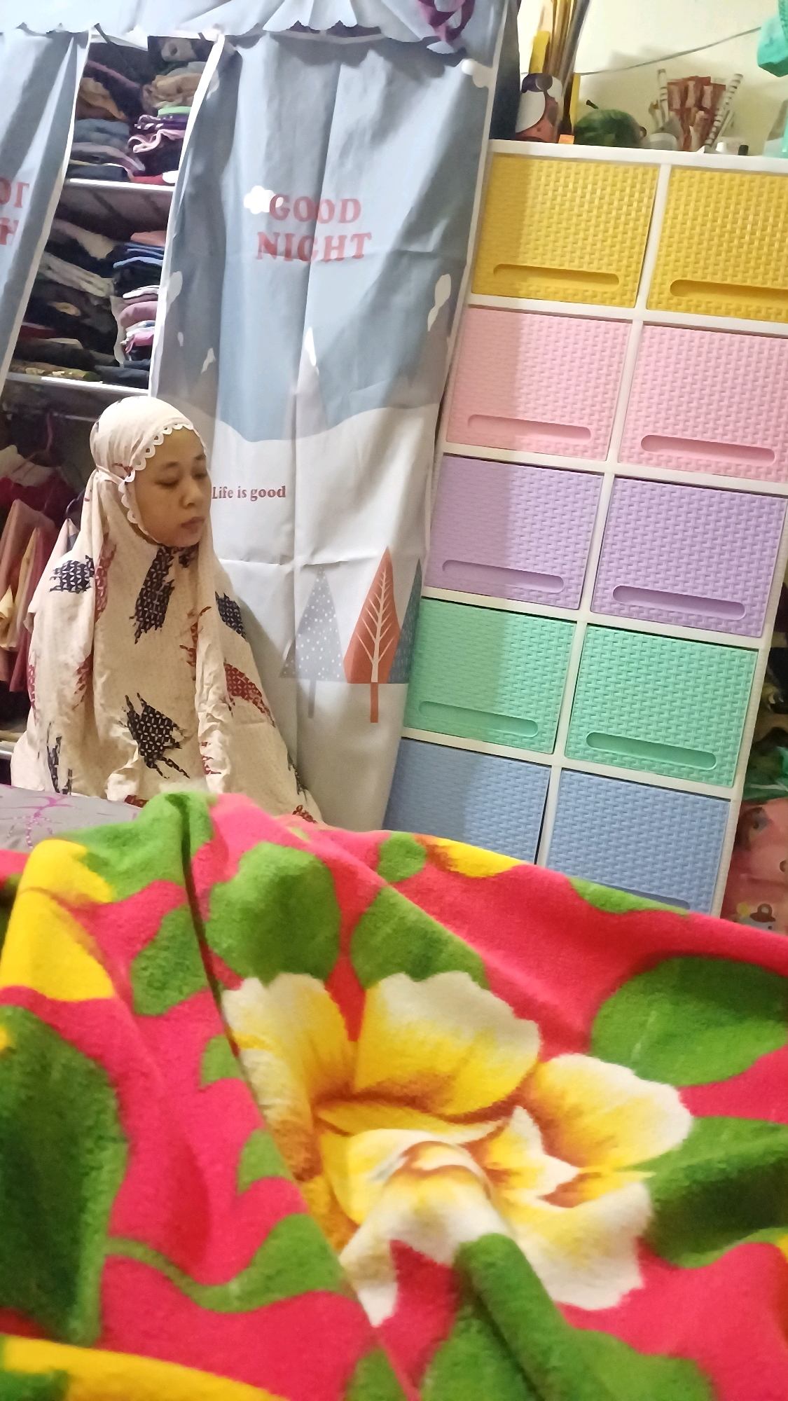 In Wonokerso, Indonesia, a woman is seen wearing a light-colored hijab and a patterned garment, possibly a prayer outfit, as she sits in front of a blue curtain with "GOOD NIGHT" printed on it. To her right, a colorful plastic cabinet with multiple drawers in yellow, pink, purple, and green is visible. The background shows a closet filled with folded clothes. In the foreground, a blanket with a vibrant floral pattern of pink, green, and yellow flowers is draped. The scene appears to be indoors, within a residential setting.