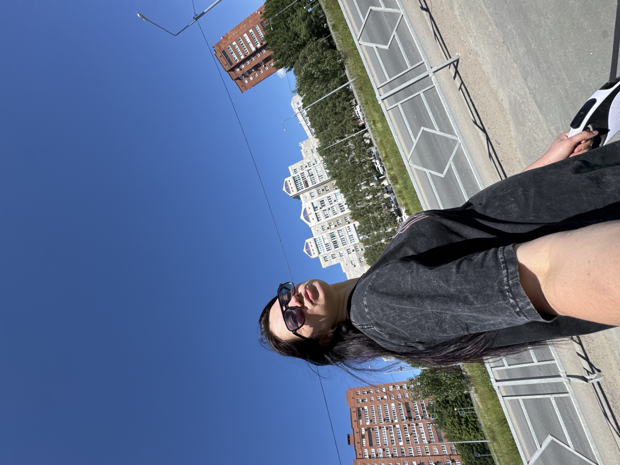 The image depicts a bright, sunny outdoor scene in an urban or suburban setting, captured in daylight. The main subject is a person, likely female, positioned on the right side of the frame. She has long, dark hair and is wearing sunglasses pushed up onto her head, looking upwards and away from the camera. She is dressed in a dark, possibly charcoal-colored, short-sleeved t-shirt with a slightly faded or textured appearance. In her right hand, visible towards the far right, she holds a white sneaker with black details.
The background features a clear, deep blue sky dominating the upper half of the image. Below the skyline, several multi-story buildings are visible, including distinctive white or light-colored residential structures, one with a notable arched architectural element, and a prominent brick-red building with many windows. Green trees are scattered among the buildings, indicating urban landscaping.
In the mid-ground and foreground, there's a paved surface, resembling a road or a wide pedestrian path, marked with geometric patterns, possibly for parking or pedestrian use. A metal fence or barrier runs alongside this paved area. Overhead, utility lines and a streetlight are visible against the sky. The strong sunlight casts crisp shadows from the fence onto the ground, suggesting a midday or early afternoon time. No discernible text is visible anywhere in the image. The overall impression is one of a casual, bright day outdoors.