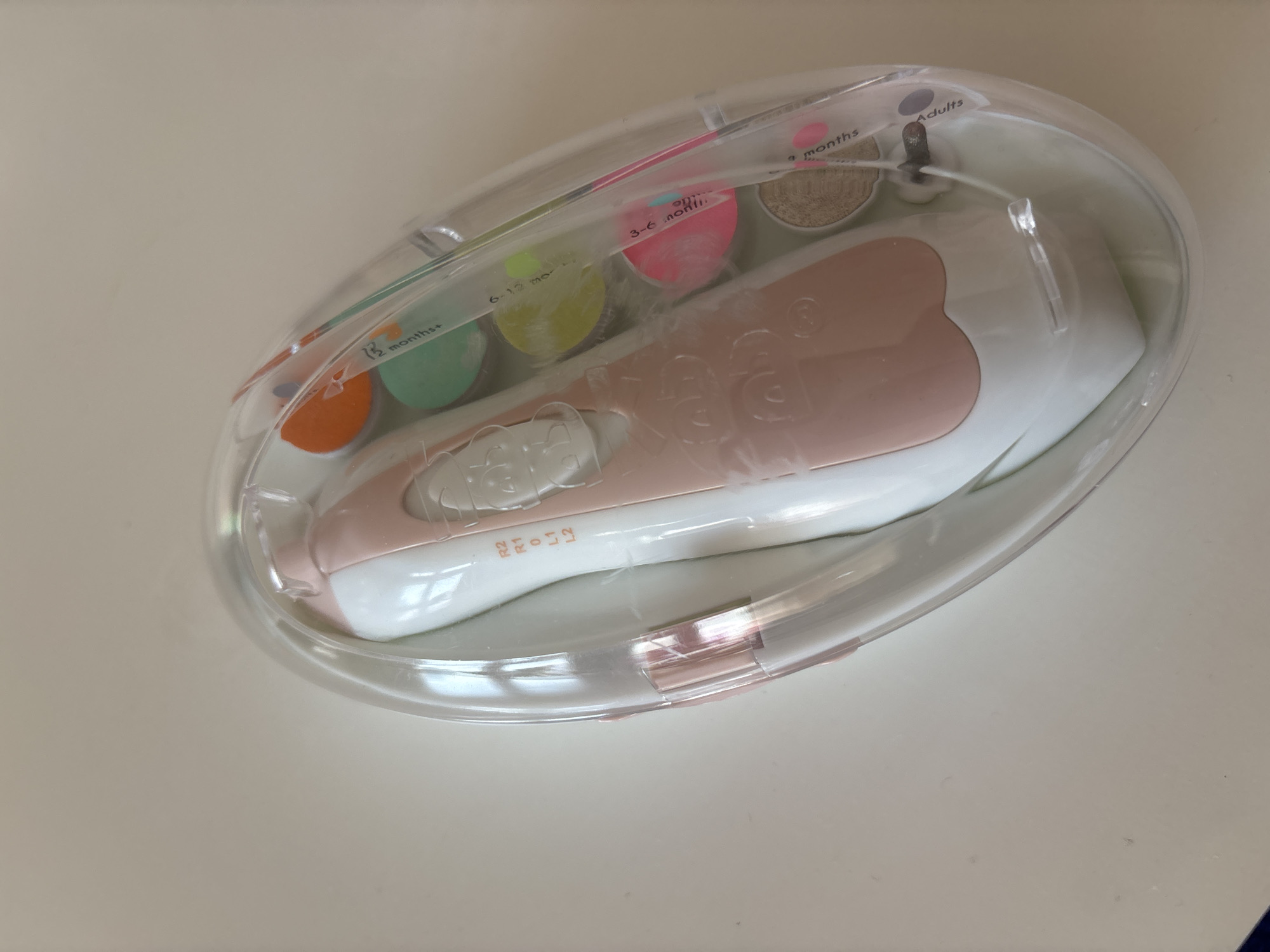 The image shows a pink and white electric nail file set inside a clear plastic oval case. The case contains the nail file, which has a pink grip and white body, along with several colored filing heads, each labeled with a different age range: "3 months+", "6-12 months", "12 months+", "18 months+", "3-6 months", and "Adults". The case is placed on a light-colored surface, likely a table. The image does not provide any information about the location of Dubai, United Arab Emirates.
