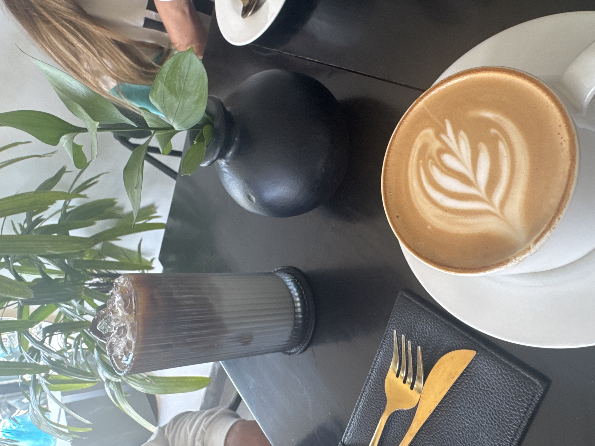 This image provides an overhead, slightly angled view of a table in what appears to be a cafe or restaurant setting. The scene features two beverages, a plant, and cutlery, suggesting a relaxed dining or coffee break.
On the right side of the frame, a white ceramic cup and saucer hold a latte with distinct, elaborate latte art resembling a leaf. To its left, a tall, ribbed glass contains an iced beverage, likely iced coffee, showing a clear separation between a dark lower layer and a lighter, perhaps milky, upper layer with ice cubes. In the center, a matte black, spherical vase holds several sprigs of a green leafy plant, contributing a natural touch to the table arrangement.
Below the iced drink, a set of gold-colored cutlery – a fork and a knife – rests on a small, black, textured mat or coaster. The table surface is dark, reflecting some ambient light. Parts of individuals are visible: long, light brown hair belonging to a person is seen in the upper left background, and an arm is partially visible in the lower left foreground, indicating people are seated around the table. The lighting is bright and soft, suggesting an indoor daytime setting. There is no visible text in the image.