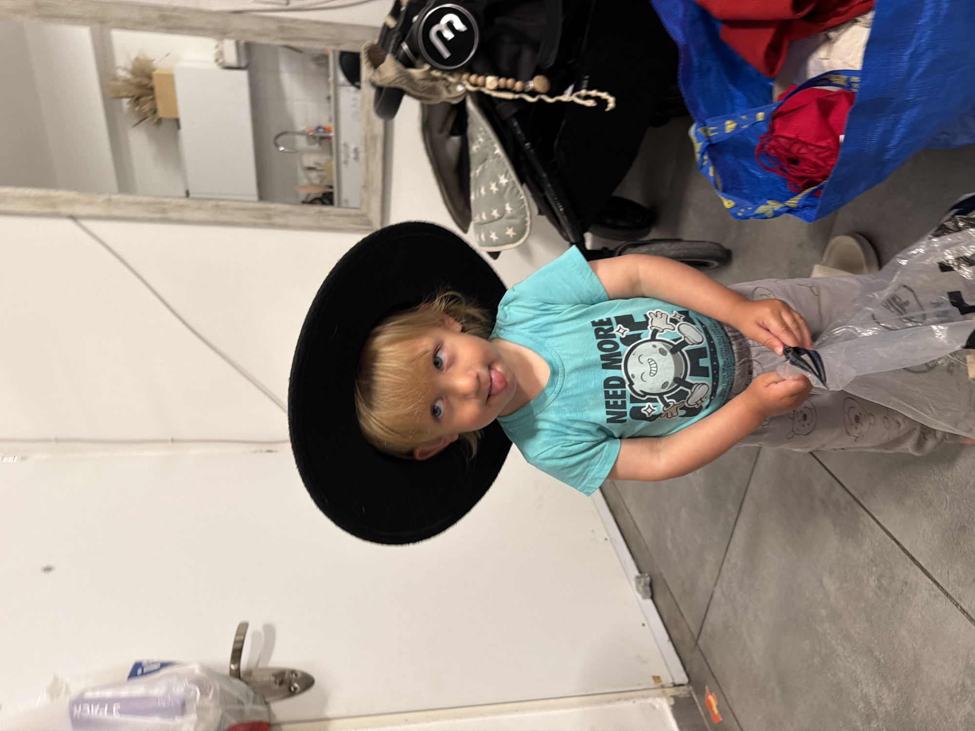The image features a young child, likely a toddler or preschooler, standing indoors. The child has blonde hair and blue eyes, and is wearing an oversized black hat and a light blue t-shirt. They are looking directly at the camera with a playful, silly expression, their tongue slightly sticking out. The child is holding a clear plastic bag in their hands.
The light blue t-shirt features a graphic of a cartoon moon or planet with a grumpy expression, and the visible text "NEED MORE". In the background, a large mirror with a light-colored frame reflects an adjacent room, revealing a sink, white appliances, and a general kitchen or utility area.
To the right of the child, a black stroller or car seat is present, featuring a grey and white star-patterned section and a visible "M" logo in a circle on its frame. Nearby, a large blue IKEA bag, filled with red fabric items, rests on the floor. The floor throughout the indoor setting is covered with large gray tiles. On the left, a white wall or door features a metal hook, with what appears to be another clear plastic bag hanging from it, showing the partial text "PACK". The scene suggests a casual domestic environment, well-lit during what appears to be daytime in Bat Yam, Israel.