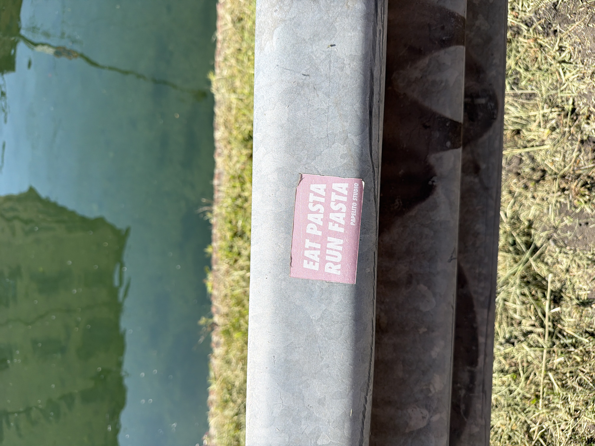 This is an outdoor photograph featuring a metal railing. Attached to the railing is a pink rectangular sticker with the text "EAT PASTA RUN FASTA" and "PAPELITO STUDIO" printed on it. To the left of the railing is a body of water, possibly a canal or river. To the right, there is grass and dirt. The location of the photo is Milan, Italy.