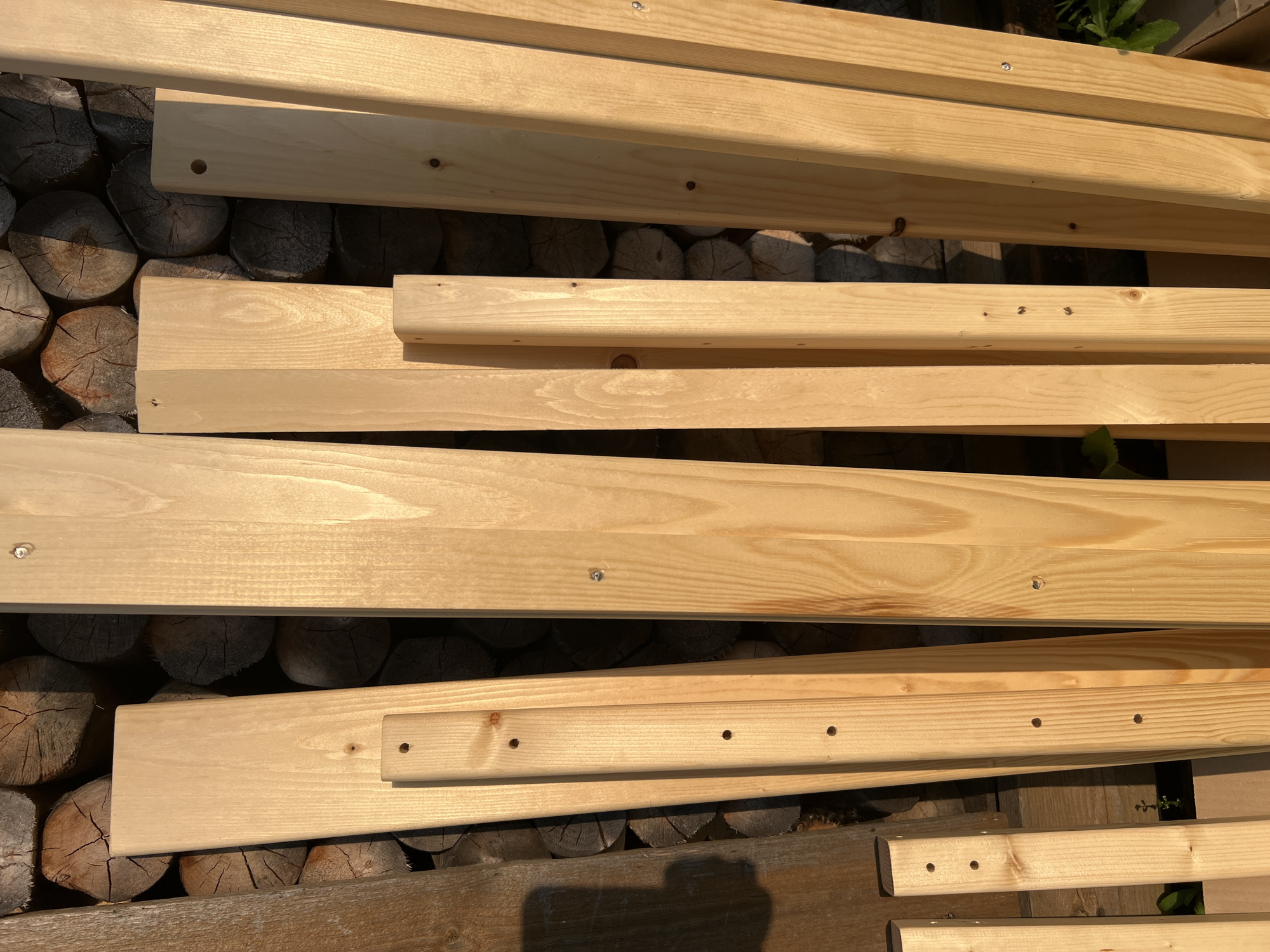 The image depicts a collection of wooden planks leaning against a stack of logs. The planks are of various widths and lengths, with some exhibiting visible knots and grain patterns. Several planks have pre-drilled holes, suggesting they are intended for construction or assembly. The scene is outdoors, as indicated by the natural lighting and the presence of some greenery in the lower right corner. The background consists of roughly cut logs arranged in a stacked fashion. The overall impression is one of a storage area for lumber, possibly associated with a rural or semi-rural setting. The location of Mochishche, Russia, is noted as context for this scene.