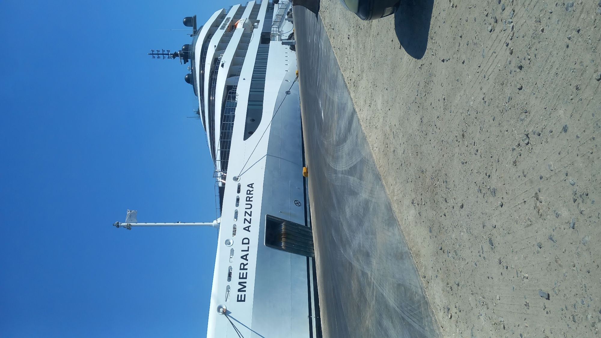 A large white luxury yacht named "EMERALD AZZURRA" is docked at a harbor. The yacht has multiple decks and a sleek design. In the foreground, a paved walkway leads away from the yacht, with tire marks visible on the surface. The ground next to the walkway is covered in sand and small stones. The sky above is clear and blue. The image appears to have been taken on a sunny day. The location is the city of Kos, Greece.