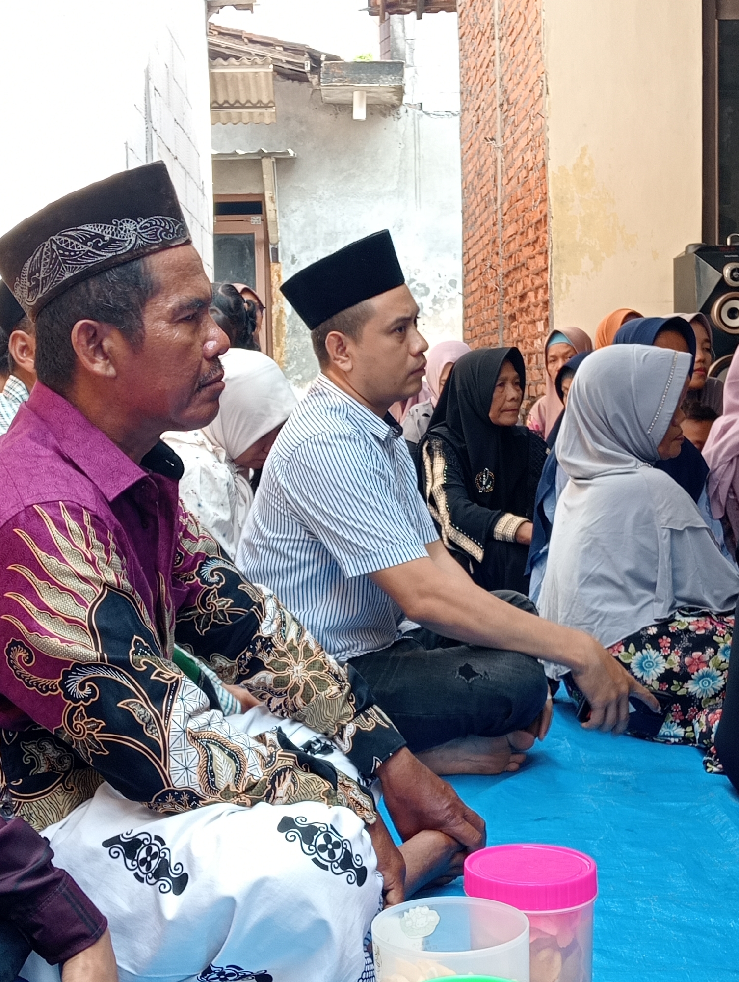 The image shows a gathering of people sitting on a blue tarp outdoors, likely in a residential area of Boyolali, Indonesia. In the foreground, two men are seated, both wearing traditional Islamic skullcaps. The man on the left wears a purple batik shirt with intricate gold and black patterns, and a white sarong with black motifs. The man next to him wears a blue and white striped shirt and dark pants. Behind them, several women are visible, mostly wearing hijabs in various colors, including black, grey, and light blue. Some are looking forward, while others appear to be looking down or conversing. The background includes a brick wall and a building with a corrugated roof, characteristic of Indonesian architecture. Two pink and clear plastic containers are placed on the tarp in the foreground. The overall scene suggests a community event or religious gathering.
