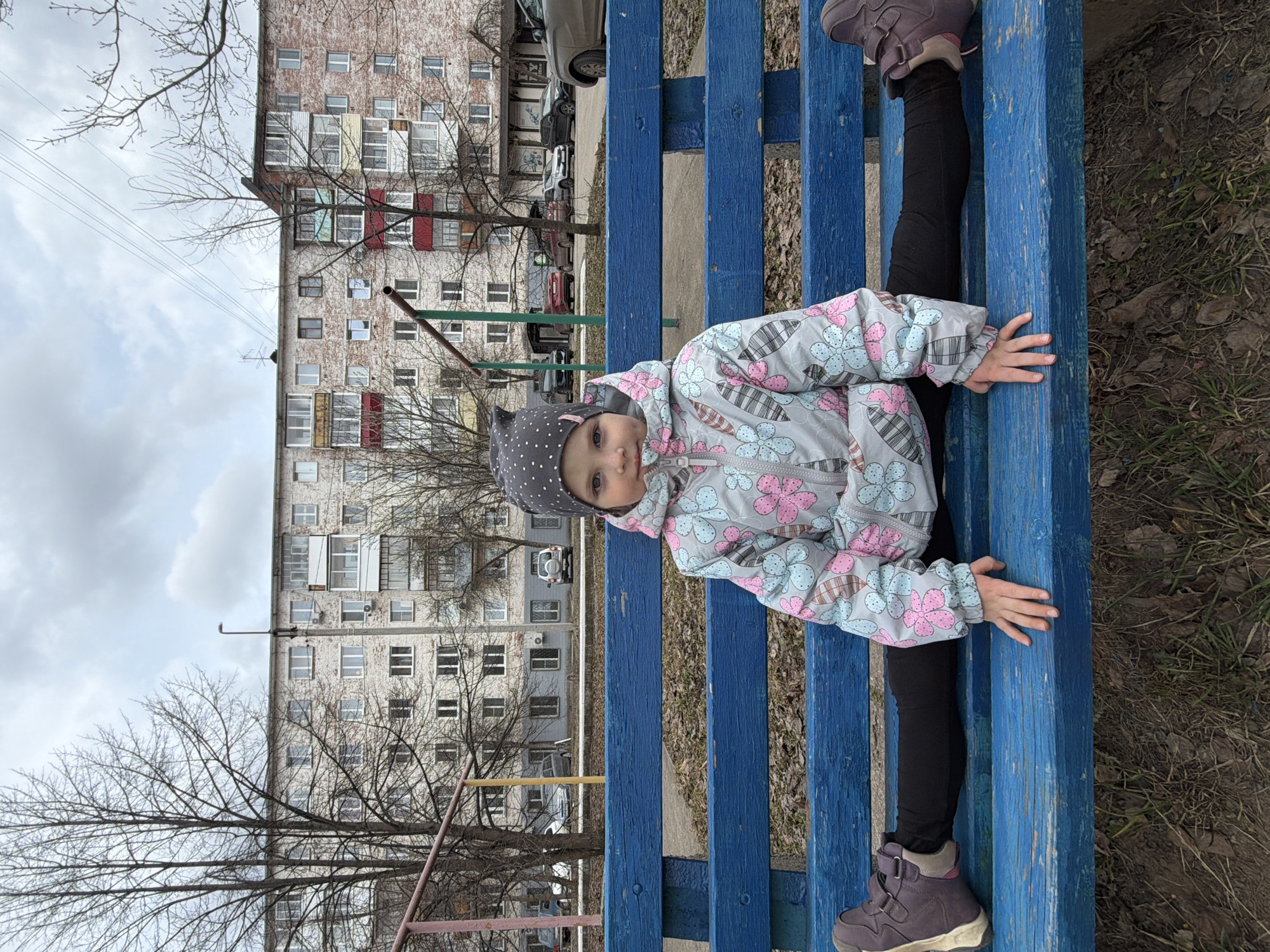 A young child sits in a split position on a blue bench. The child wears a gray and pink floral-patterned jacket and a gray, polka-dotted hat. The child is facing the camera with a neutral expression. The background includes an apartment building, bare trees, and a cloudy sky. The location is likely in Votkinsk, Russia, given the prompt.