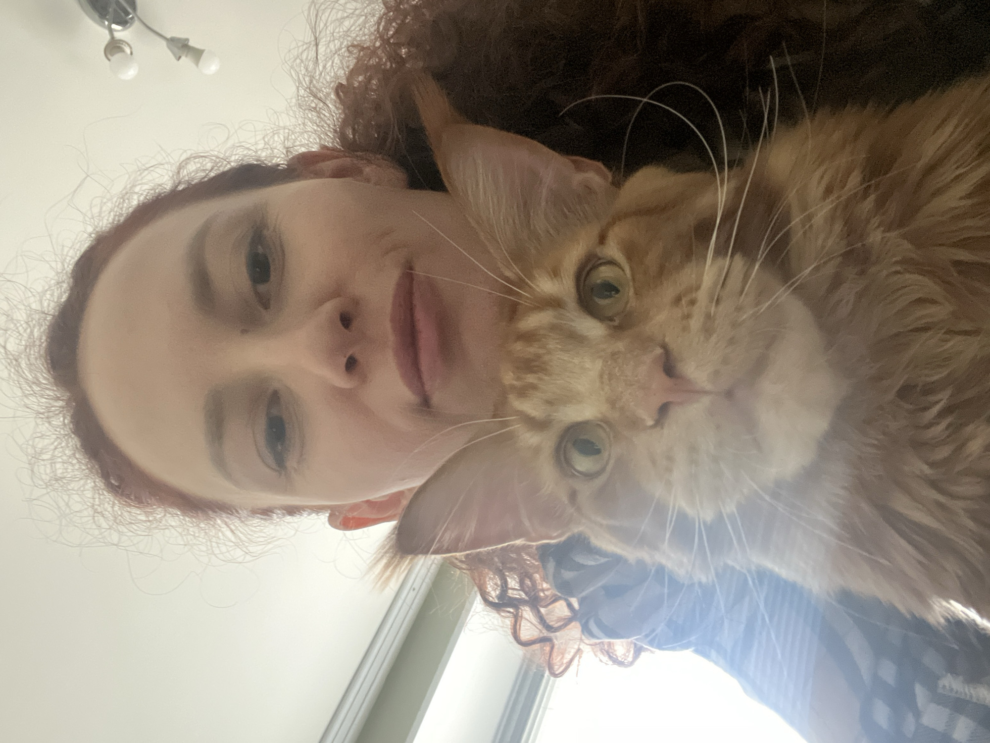 A selfie-style, close-up photograph depicts a red-haired woman and a ginger cat. The woman's face is visible with fair skin, curly red hair, and neutral facial expression. The cat, a large ginger breed possibly a Maine Coon, is positioned near her face, partially obscuring her. The background consists of a white ceiling and a window with light coming through. The location is unknown and is not in the city of Saint Petersburg, Russia.