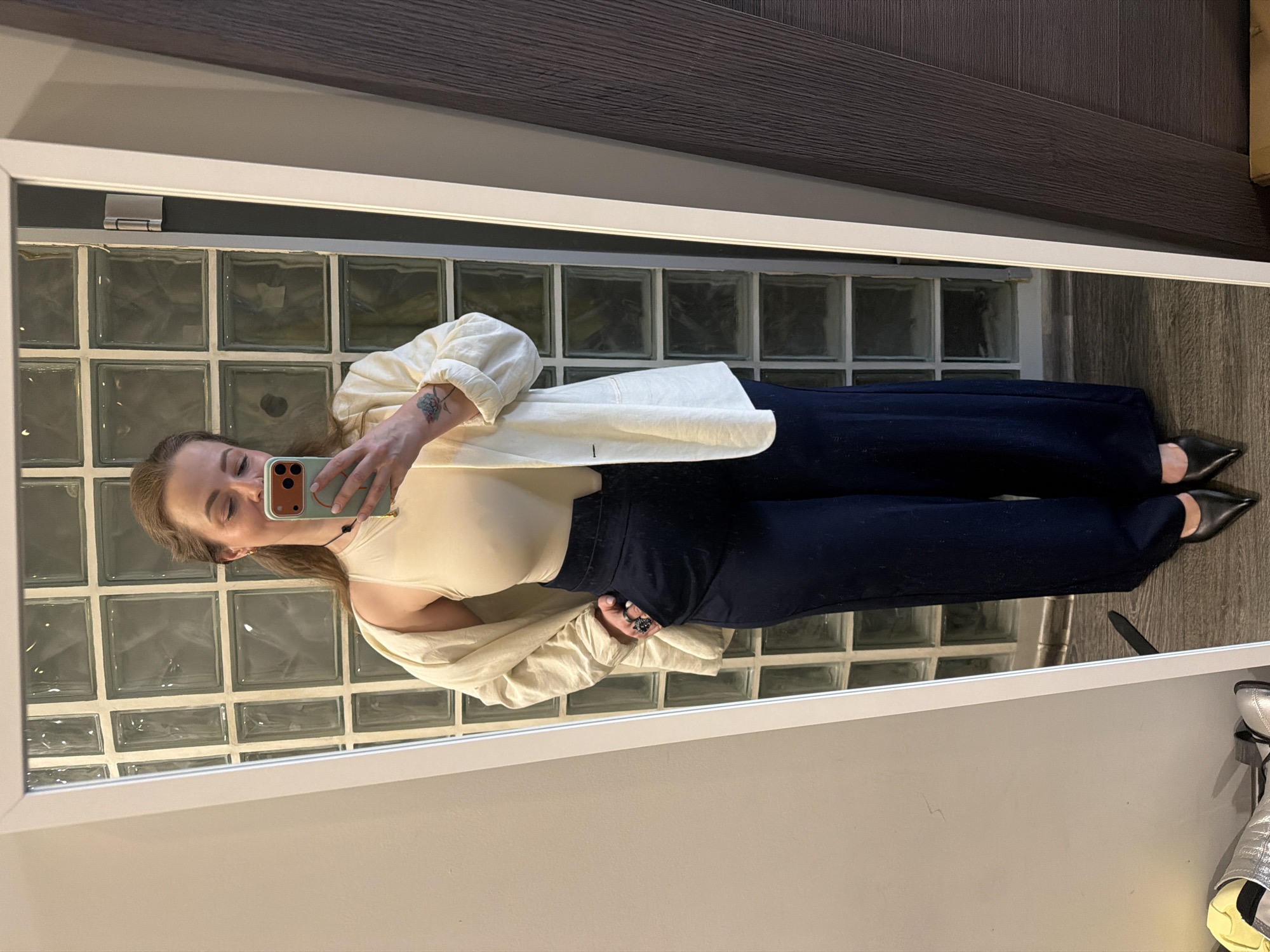 A woman is captured in a mirror selfie, standing upright and holding a light green smartphone with an orange camera module to take the picture. She has light brown hair, possibly pulled back, and her eyes are cast downwards or closed. She wears a cream or beige sleeveless top, an open, long-sleeved light off-white shirt, and dark navy wide-leg trousers. Black pointed-toe shoes are visible on her feet. A small, delicate floral or butterfly tattoo is visible on her left forearm/wrist, and she has a ring on her right hand.
The setting, reflected in the large white-framed mirror, appears to be an indoor space in Moscow, Russia. Behind her, a wall composed of square glass blocks provides a textured, translucent background. Above the mirror, dark, textured wooden paneling extends across the wall or ceiling, contrasting with a thin, linear light fixture mounted just above the mirror's frame. A silver hinge is visible on the upper left side of the mirror's frame. In the bottom right corner of the reflection, part of a metallic silver shoe or boot can be seen, hinting at a retail or dressing room environment. No discernible text is visible in the image.