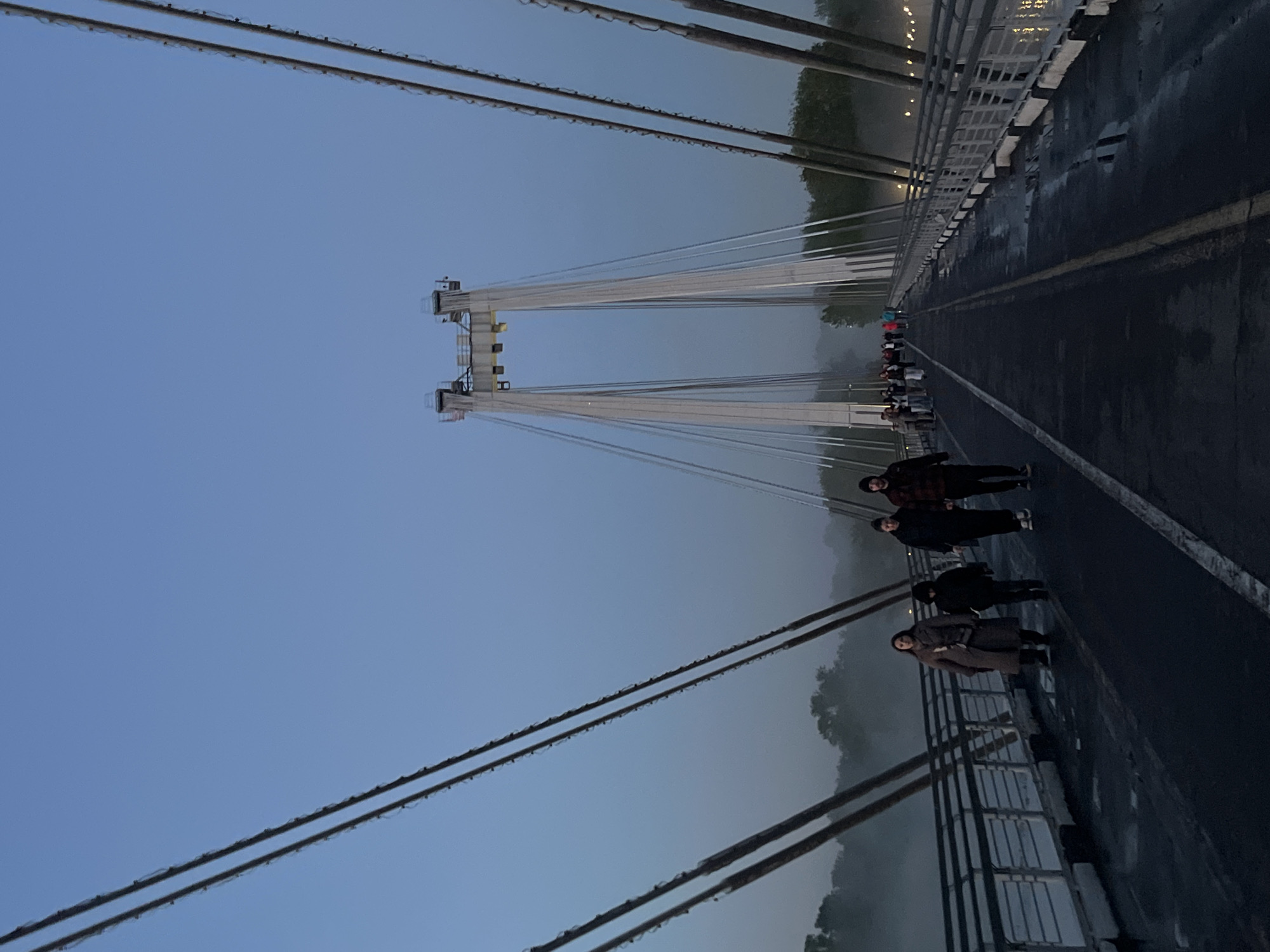 The image depicts a group of people walking on the Vinogradovsky Bridge in Krasnoyarsk, Russia. The bridge is a cable-stayed bridge, and its towers and cables are prominently visible. The sky is overcast and the atmosphere is foggy or misty. The bridge deck is wet, likely from rain or fog. Several people are walking along the bridge. They are dressed in casual clothing. In the distance, more people can be seen. The bridge railings are visible on both sides of the walkway. Trees on the banks of the river are partially obscured by the fog.