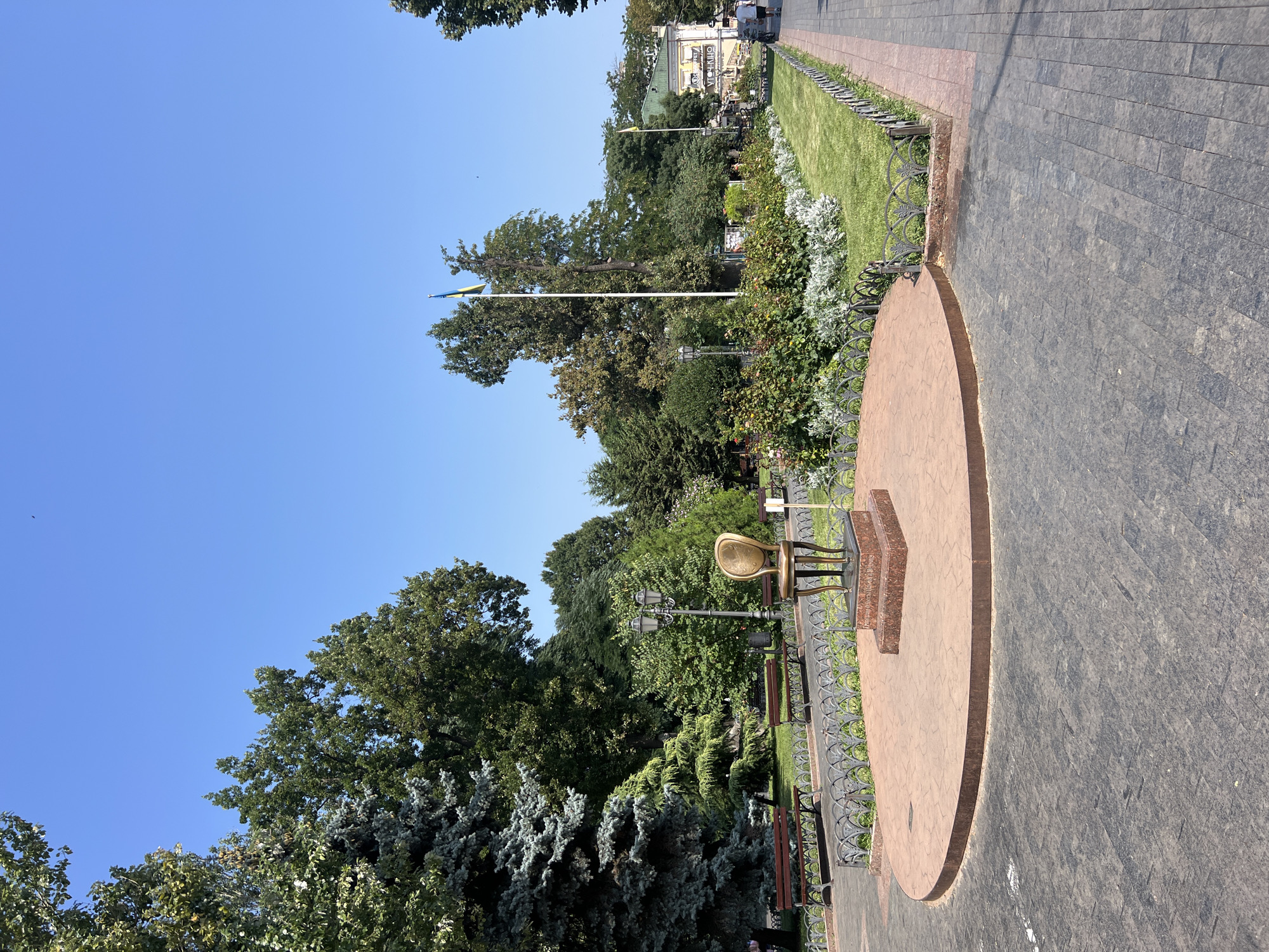 This is a medium-shot, eye-level, outdoor, daytime, full-color photograph of the "Throne of the Twelve Chairs" monument in Odesa, Ukraine, located in the City Garden. The monument, a golden chair with intricate detailing, sits atop a circular brick platform in the foreground. The platform is surrounded by a decorative metal fence. Beyond the fence is a lush green lawn with flower beds and trees. In the background, two Ukrainian flags fly from tall poles, and a building with a green roof is visible. The ground in the foreground and along the pathway is paved with dark gray and red bricks. A few people are walking along the pathway in the background. The sky is clear and blue.