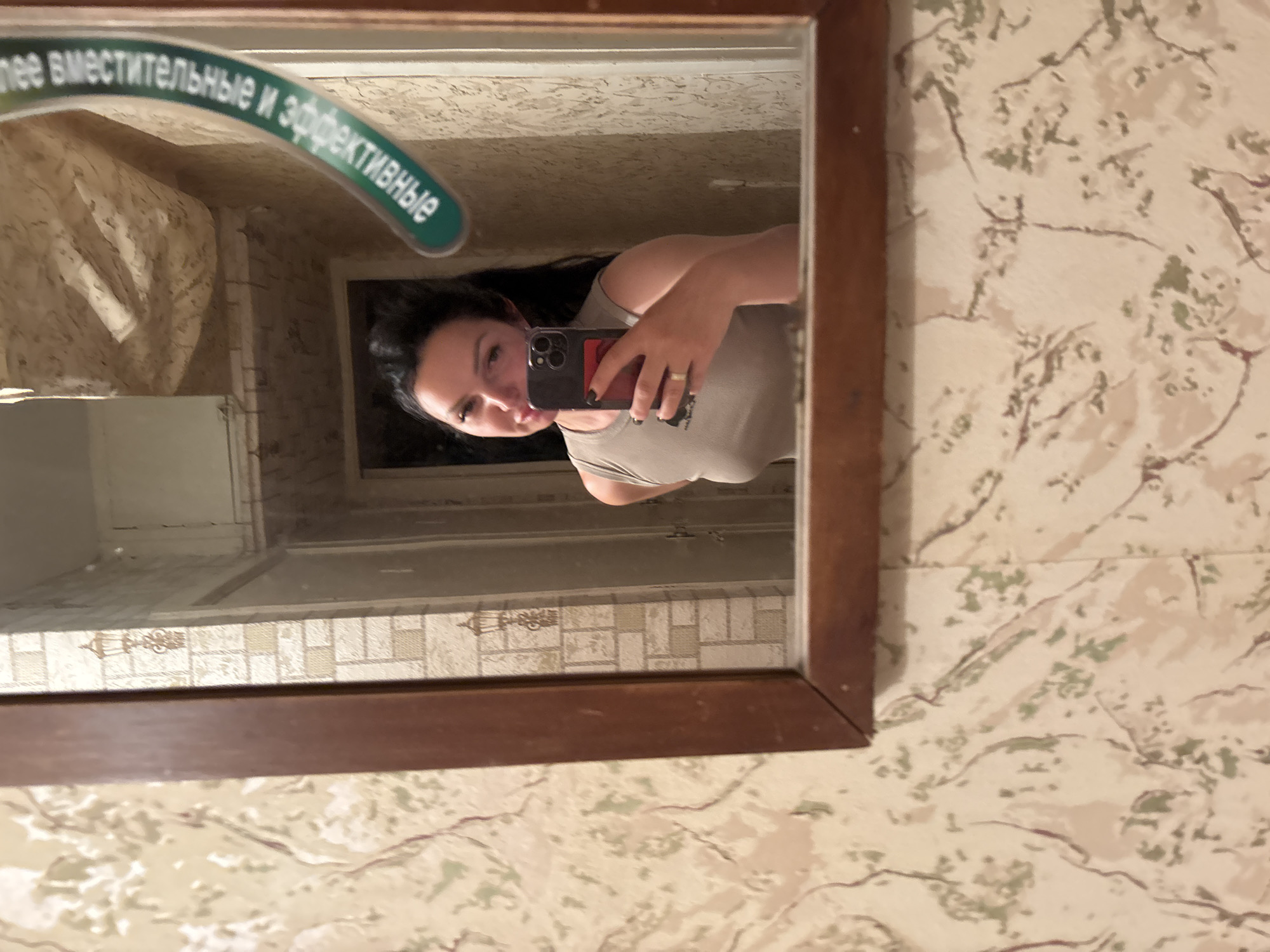 A selfie of a woman reflected in a mirror. The woman is holding a smartphone and looking at the camera. The mirror is framed with dark brown wood. The wall behind the mirror is decorated with floral-patterned wallpaper. Above the mirror, a curved sign displays text in Russian: "лее вместительные и эффективные." The scene appears to be an interior setting, likely a bathroom. The presence of the Russian text suggests a location within Russia, possibly Votkinsk.