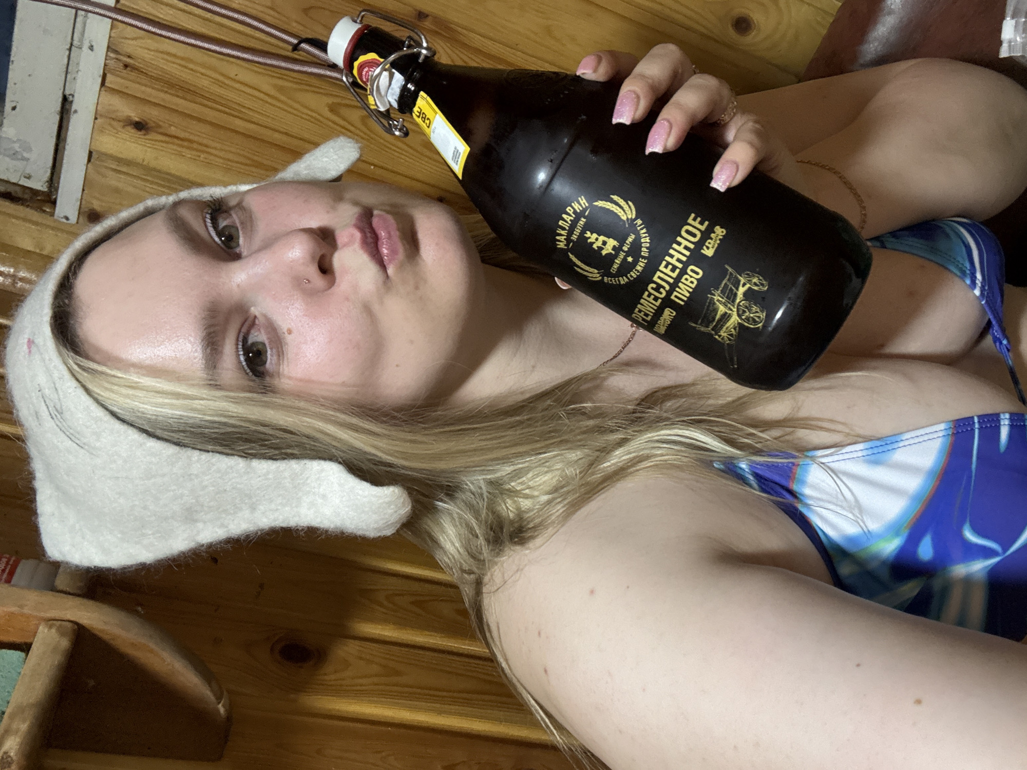 A woman with fair skin and blonde hair, wearing a white sauna hat and a blue and white swimsuit, is holding a dark-colored bottle of beer. The bottle has a label written in Russian, stating the beer is from the "Maklarin Brewery" and "Craft Beer" ("РЕ МЕСЛЕННОЕ ПИВО"). The background appears to be a wood-paneled sauna. A partial view of a wooden bench is visible in the bottom left corner.
There is no information about the city of Dubovaya, Russia in the image.