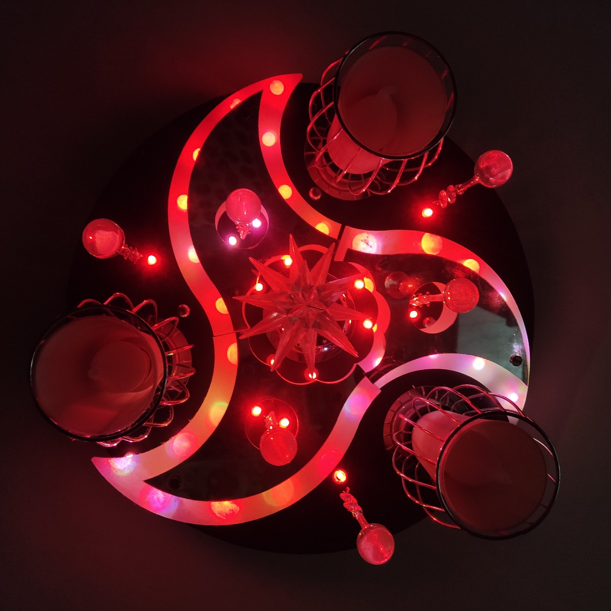 A high-angle shot shows a round, dark table surface illuminated with red and orange lights in a spiral pattern. At the center of the table is a multi-pointed star decoration made of clear material. Three metal-framed cups or containers are arranged in a triangular formation around the center. Between each cup, there's a small, spherical object with an ornate handle, also illuminated with red light. The background is dark and out of focus, suggesting a dimly lit room. The scene captures a decorative arrangement, possibly for a festive or celebratory event. I have no information about its location in Krasnodar, Russia.