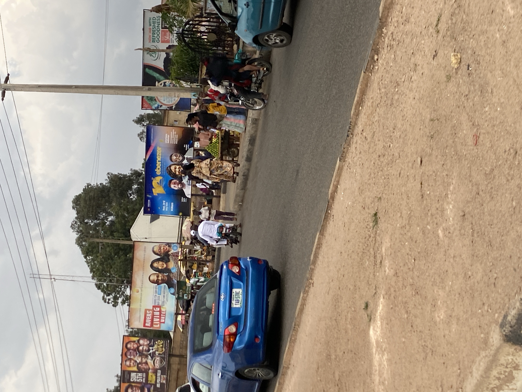 The image depicts a street scene in what appears to be Gura Top, Nigeria, with various vehicles and people present. A blue sedan, with license plate YAB-57KC, is prominently positioned in the foreground. Along the roadside are multiple billboards advertising different products and services, including "RIVERS LIVING WATER" and an "ebenezer" advertisement. People are walking along the road and riding motorcycles. The setting includes telephone poles with wires and a partly cloudy sky. The overall environment suggests an urban or semi-urban location with active street life.