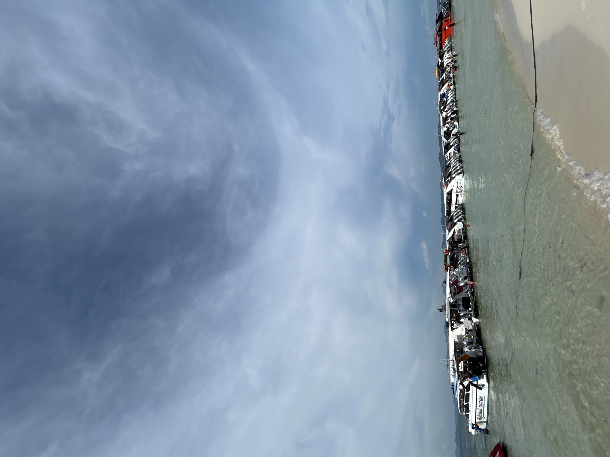 The image shows a view of a tropical beach in Tambon Phru Nai, Thailand. The foreground features a sandy beach with a gentle wave lapping the shore, and a rope anchoring a series of speedboats. The clear, shallow water reveals the sandy seabed. A long line of speedboats, predominantly white with some red accents, are moored parallel to the shore, suggesting a popular tourist destination. Several people are visible on the boats and in the water, some appearing to be disembarking or preparing to board. The background consists of a hazy, mountainous coastline under a partly cloudy, grey sky. The overall scene depicts a bustling marine activity area.