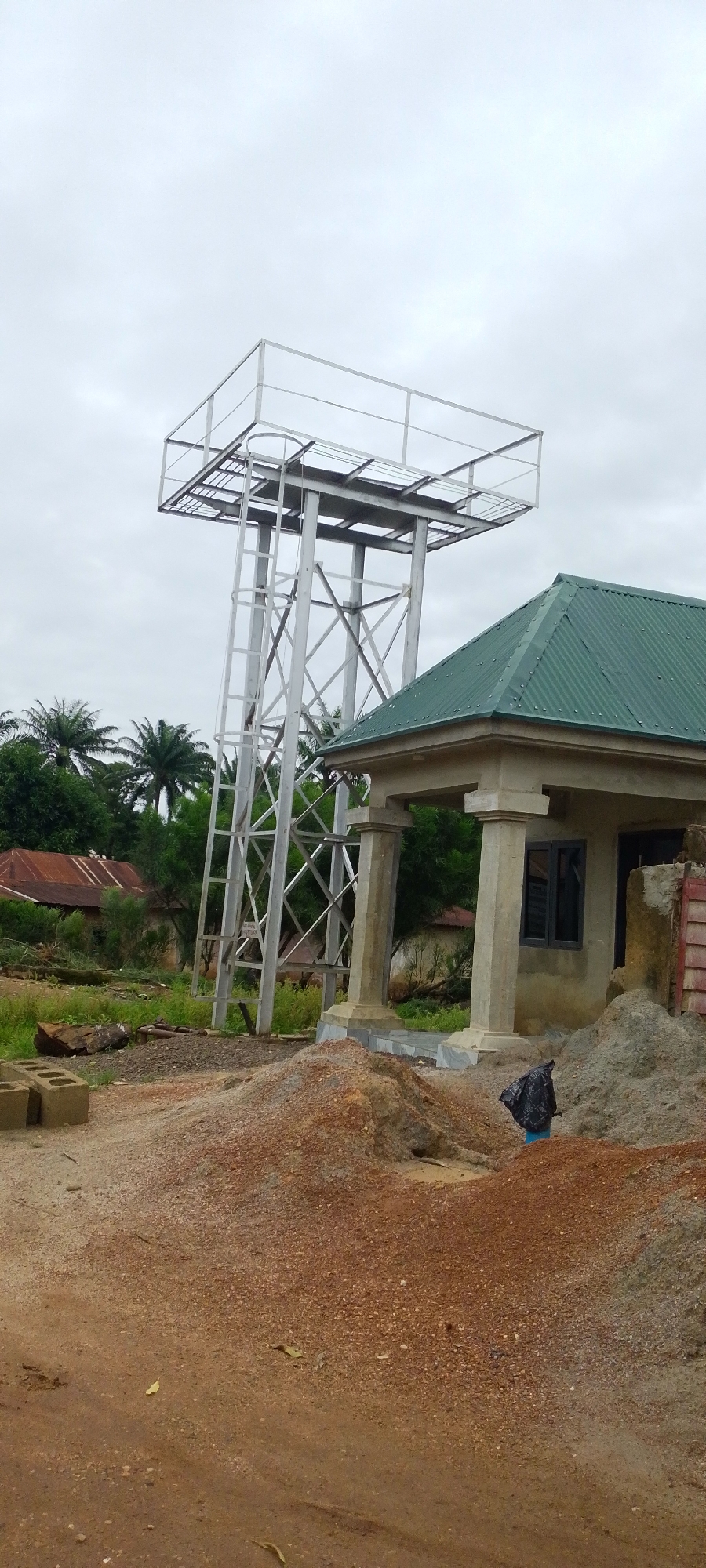 In the city of Baissa, Nigeria, a partially constructed building with a green corrugated metal roof stands beside a tall, silver metal water tower. The water tower has a square platform at the top with a metal railing and a ladder running up its side. The building features a covered porch with two large, square concrete pillars. In the foreground, there are piles of red and grey gravel and sand, as well as some scattered construction materials like cinder blocks. The background reveals a lush green landscape with palm trees and other vegetation, along with a few houses with corrugated metal roofs. The sky is overcast. A black plastic bag is visible near the building.