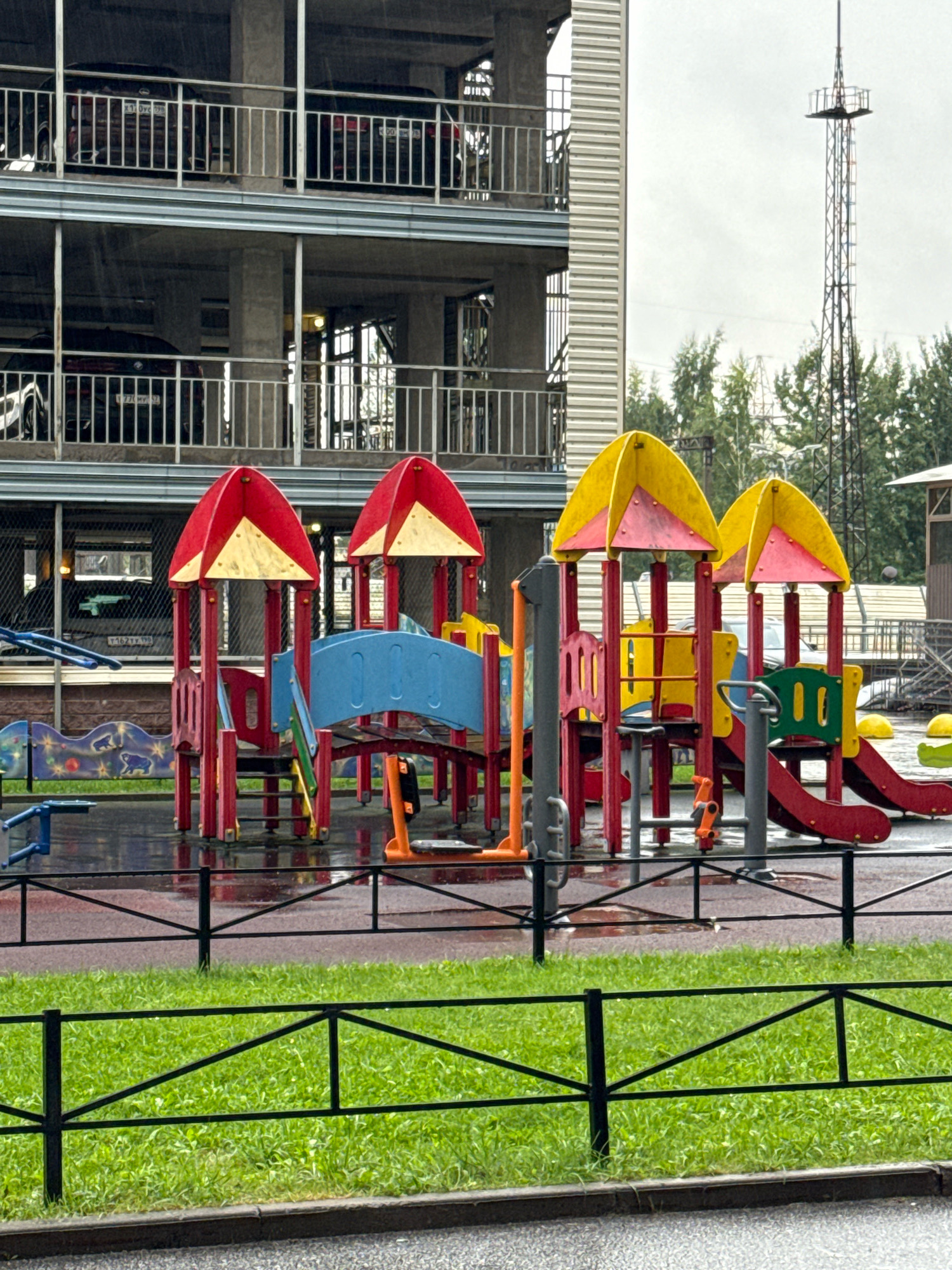 The image captures a playground situated next to a multi-story parking garage, with a tall tower visible in the background. The playground equipment is colorful, featuring red and yellow canopies, blue and red slides, and various climbing structures. The ground of the playground appears wet, suggesting recent rain. A black metal fence separates the playground from a strip of green grass and a concrete walkway in the foreground. Cars are visible on multiple levels of the parking garage, and trees can be seen beyond the garage. The overall scene suggests an urban environment in Russia, possibly on a cloudy day given the lighting. No people are present in the image.