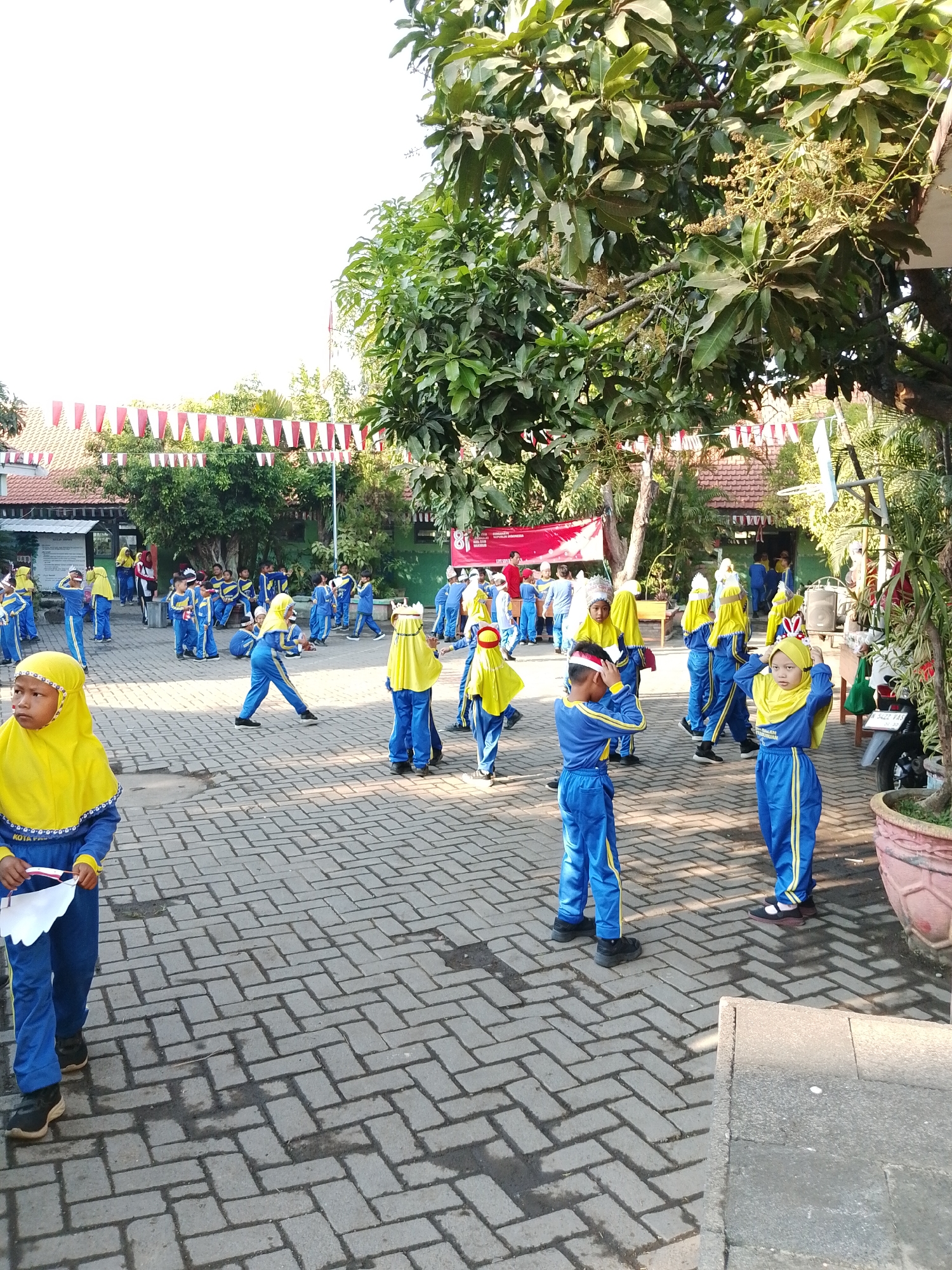 The image shows a group of young children, likely students, dressed in bright blue uniforms with yellow headscarves and trim, gathered in an outdoor paved area. They appear to be participating in some kind of activity, possibly a school event or performance, given their coordinated outfits and the celebratory decorations. Red and white triangular flags are strung across the background, suggesting a national holiday or celebration. A banner with Indonesian text is visible, and some children are holding white papers. The environment includes trees with green foliage and some buildings in the background. The ground is paved with interlocking bricks. The scene suggests a lively and festive atmosphere at a school in Pasuruan, Indonesia.