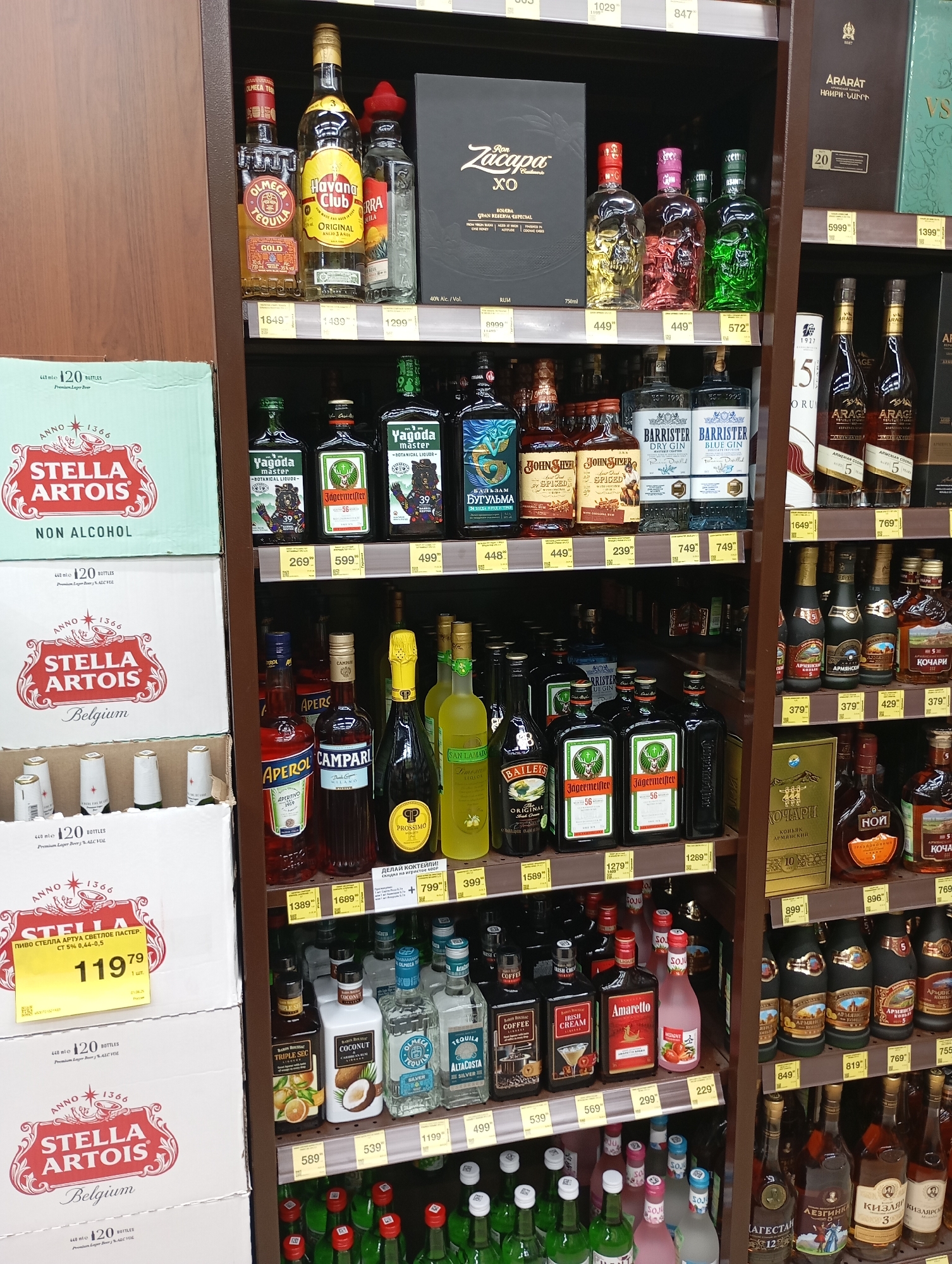 The image displays a retail shelf in a Moscow, Russia store, likely a liquor store or supermarket aisle, showcasing a variety of alcoholic and non-alcoholic beverages. The shelves are organized with numerous bottles and some boxes of drinks.
Prominent items include:
* Several bottles of Stella Artois beer, with two large boxes labeled "Stella Artois Belgium" and "Stella Artois Non Alcohol" visible on the left side.
* Various types of gin, tequila, and rum, including brands like "Barrister," "Olmeca," "Yagoda Master," and "Ron Zacapa XO."
* Other bottles such as Jägermeister, Aperol, Campari, and liqueurs like "Amaretto."
* A display of Armenian brandy.
* A variety of smaller bottles, some appearing to be liqueurs or spirits, with labels in Russian.
The lighting suggests an indoor environment, likely illuminated by store lighting. The shelves are constructed from metal and wood. Prices are visible on some of the items, indicated by yellow tags. The scene is a static product display, with no people or active interactions depicted. The overall impression is one of commercial merchandise presented for sale.