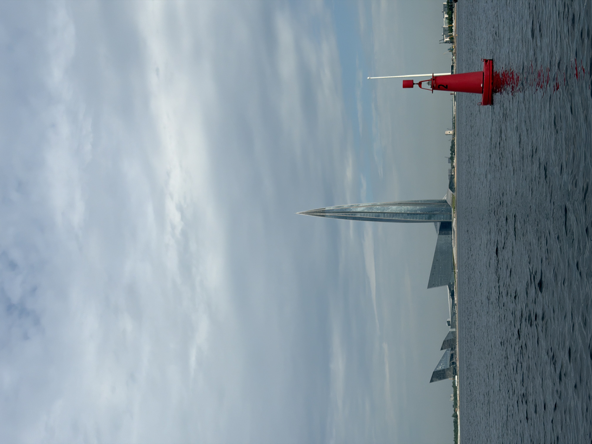 The image captures a view of the Lakhta Center skyscraper in Saint Petersburg, Russia, from the Gulf of Finland. The skyscraper, characterized by its tall, spiraling glass facade, dominates the skyline. To its left are other modern architectural structures, also appearing to be part of the Lakhta Center complex, with angular, glass-covered designs. In the foreground, the dark, rippling waters of the Gulf of Finland are visible, with a bright red navigational buoy, numbered '2', floating prominently in the right side of the frame. A white pole extends vertically from the buoy. The sky is overcast with a mixture of grey clouds and patches of blue, suggesting a potentially cool or moderate day. The overall scene conveys a sense of modern urban development set against a natural waterfront environment.