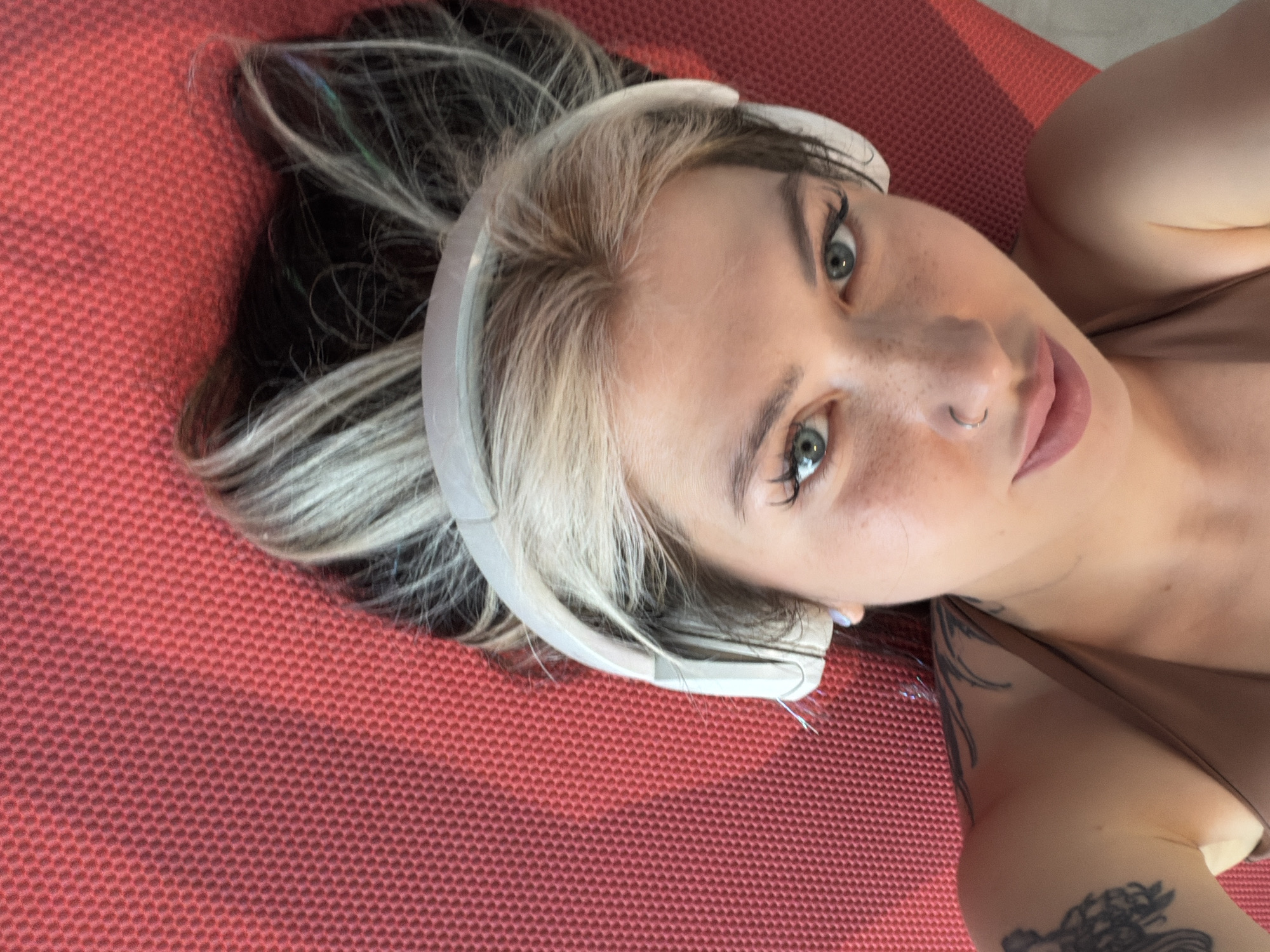 A woman with dyed blonde and black hair is lying on a red textured surface, possibly a yoga mat. She is wearing white headphones. Her face is directed toward the camera, revealing green eyes, freckles, and a nose ring. A tattoo is visible on her arm, and there is no indication this image was taken in Budva, Montenegro.