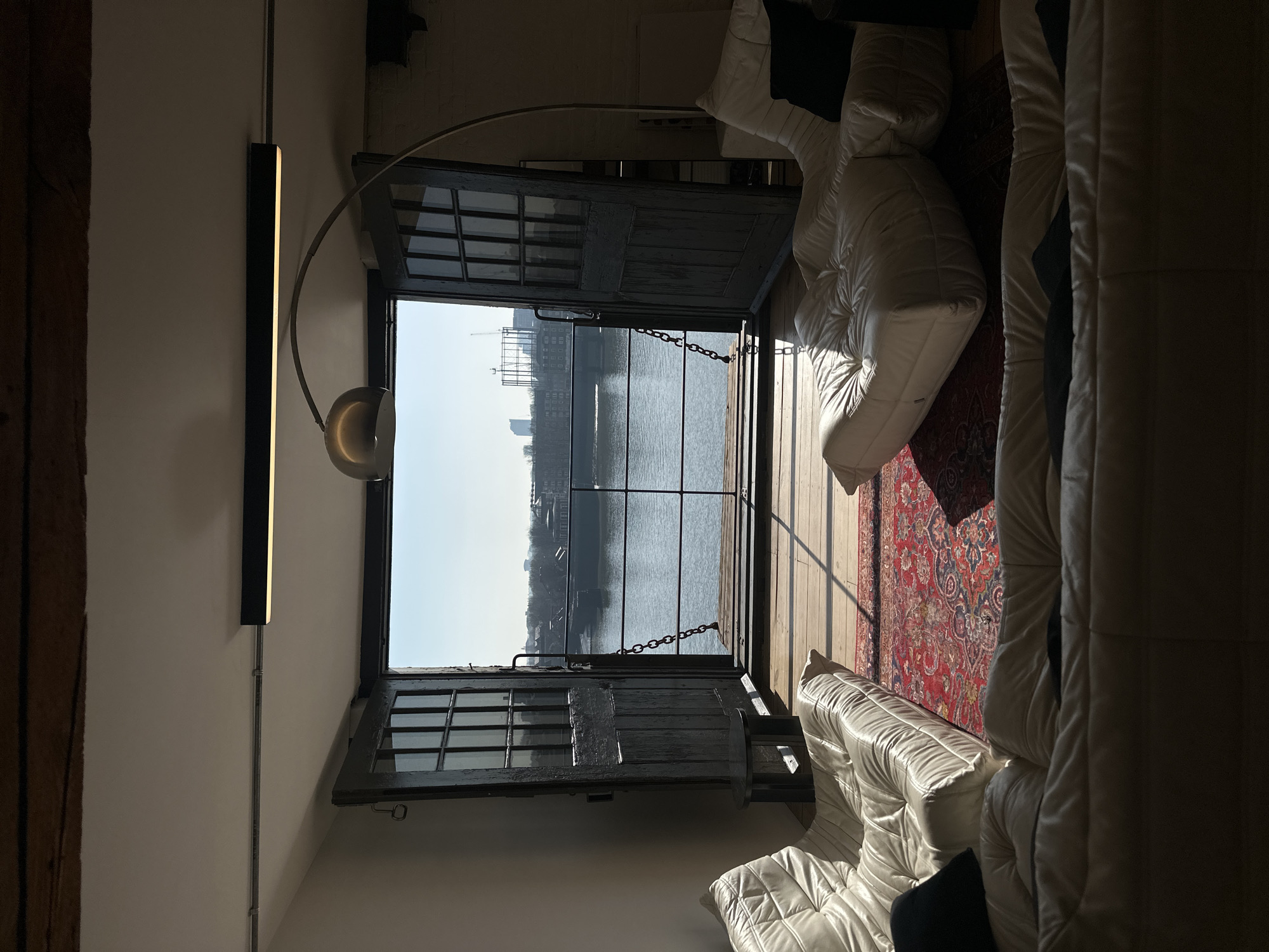 The image displays the interior of a room within a riverside apartment located in London, United Kingdom, offering a direct view of the River Thames. The room is characterized by a white ceiling with an exposed dark wooden beam and a black linear LED light fixture mounted with exposed conduit. The floor consists of light-colored wooden planks partially covered by a large, ornate red Persian-style rug.
Three distinct white or cream-colored modular sofas are arranged around the rug, facing the open exterior. Two black throw pillows are visible on separate sofas. A small, dark, round side table with a pedestal base is positioned near the left sofa. On the right side of the room, an arching metallic floor lamp with a bowl-shaped shade stands.
The room's far wall features large, dark-stained, double wooden doors, each fitted with multiple glass panes, which are open to reveal a small wooden balcony or deck area situated directly over the water. A black metal railing with horizontal bars and vertical chain segments forms a barrier along the balcony's edge.
Beyond the railing, the River Thames is visible under a clear, bright blue sky. A large, dark, flat-topped barge or industrial vessel is moored on the river. The opposite bank features lower-rise buildings, and in the distant background, a prominent skeletal gasholder frame is discernible, consistent with views towards areas like the Greenwich Peninsula. Strong sunlight streams through the open doors, casting distinct shadows within the apartment. The scene is devoid of people.
