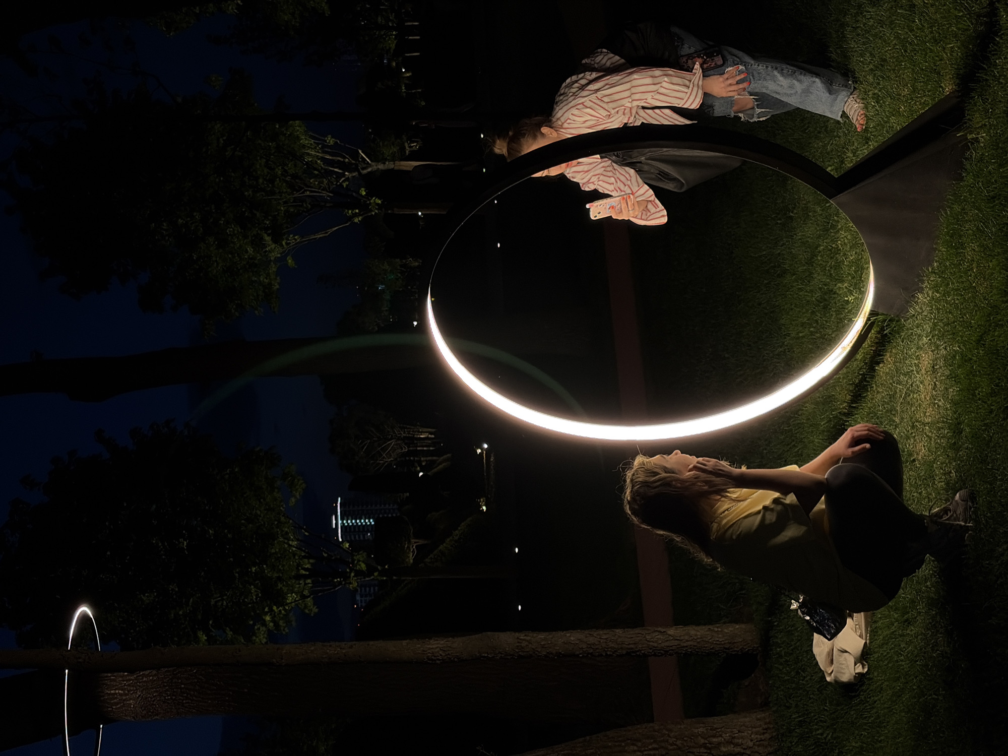 Two women are in a park at night in Krasnodar, Russia. One woman sits on the grass wearing a yellow t-shirt and black pants. A white bag is next to her. The other woman leans over a circular light fixture, with her reflection visible in the surface. She wears a red and white striped top and ripped jeans. The park is dark, with trees in the background and a building illuminated in the distance. Another smaller light fixture is visible in the background.
