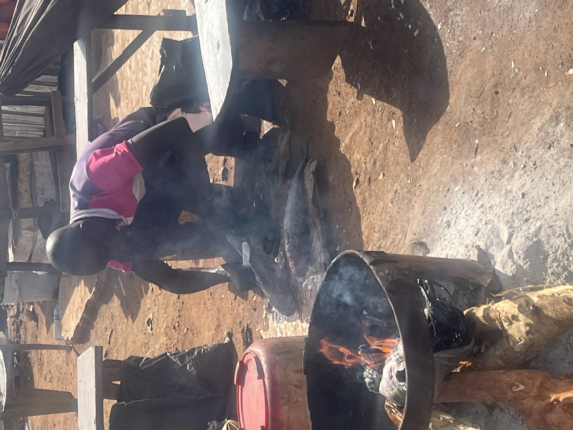 This image captures an outdoor scene in Gura Top, Nigeria, featuring a person engaged in an activity involving fire and cooking or preparation.
The main subject is a person, likely a young individual or child, dressed in a long-sleeved shirt that is purple and pink, with dark trousers. Their face is partially obscured as they lean over a structure, appearing to interact with something beneath it. Their head is visible, turned towards the ground.
The scene takes place on what appears to be bare, dusty ground, typical of an outdoor, possibly rural or semi-urban setting. Prominent shadows suggest bright sunlight, indicating it is daytime. The ground is uneven and light brown.
Several objects are visible. In the foreground, there's an open metal barrel, cut in half lengthwise, with a fire burning inside, producing smoke and flames. This setup appears to be a makeshift stove or grill. Next to this, a red plastic container (possibly a barrel or large bucket) is on the ground. Another structure, possibly made of wood and metal, is near the person. The person seems to be attending to something on or near this second structure, from which smoke also emanates, suggesting another cooking or heating point. Various wooden planks and poles form part of a crude shelter or framework in the background and around the scene. A small white bottle is visible on the ground in the mid-background.
The activity taking place is centered around open-air cooking or heating using wood fires in makeshift containers. The person is actively involved, leaning into the setup, perhaps stoking the fire, arranging items, or preparing food. The presence of smoke indicates active burning.
Notable details include the simple, functional nature of the cooking setup, the bright sun casting strong shadows, and the earthy environment. No visible text is discernible in the image.