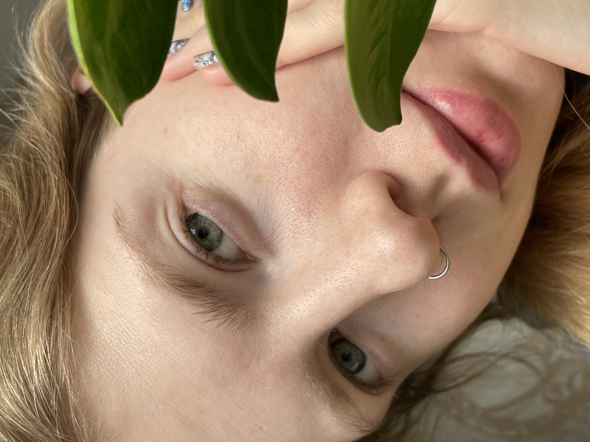 A close-up, inverted portrait captures a person with fair skin, light blonde or light brown hair, and striking light green or hazel eyes. The subject's head is tilted, giving the image an unconventional perspective. Two large, vibrant green leaves partially frame the face in the foreground, obscuring the forehead and upper cheeks, adding a natural, artistic element to the composition.
The person gazes directly towards the viewer with a calm, neutral, or contemplative expression. A silver C-shaped nose ring is visible on the septum. Their lips appear full and are colored with a soft pink or reddish hue, possibly from lip gloss or a natural tint. Light-colored eyelashes and well-defined eyebrows are visible.
A hand with painted fingernails, featuring a delicate light-colored, possibly abstract or floral, design, is subtly visible in the upper right area, resting near the hair. The skin exhibits natural texture, including visible pores and subtle variations in tone. The lighting is soft and diffused, characteristic of indoor conditions. The background is indistinct, keeping the focus entirely on the subject's face. The image depicts a posed moment rather than an activity. There is no visible text. The photograph was taken in Izhevsk, Russia.