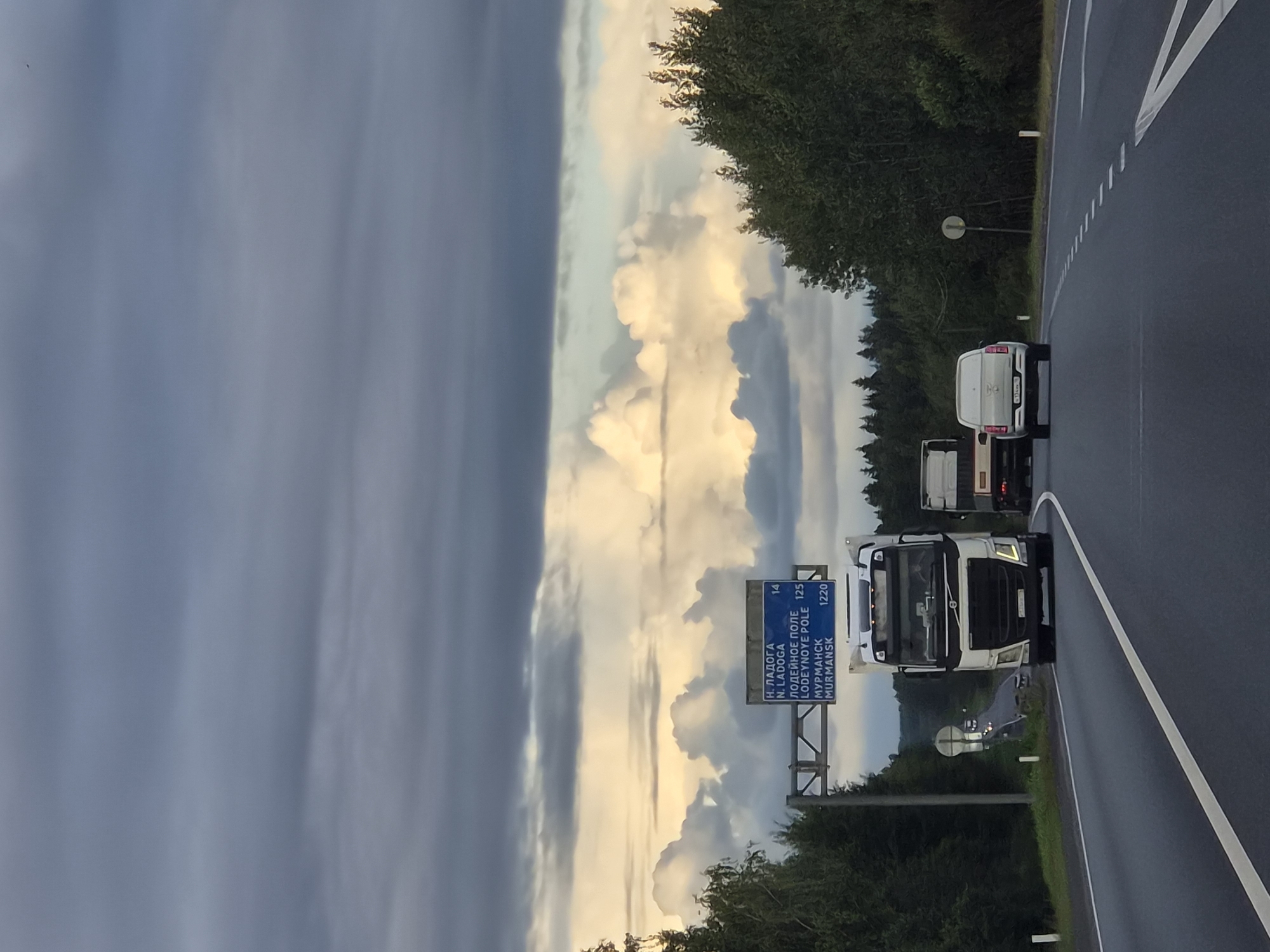 The image depicts a highway scene in Kisel'nya, Russia, under a dramatic sky filled with dark and light clouds. A white Volvo semi-truck, with a white trailer, is prominently featured in the foreground, traveling on the right lane of a multi-lane asphalt road. To the right of the Volvo, a white minivan is also traveling in the right lane. In the distance, another semi-truck is visible in the left lane. A blue highway sign is suspended over the road, displaying destinations and distances: "N. LADOGA 14", "LODEINOYE POLE 125", and "MURMANSK 1220". The roadside is lined with dense green trees. The lighting suggests it is either early morning or late afternoon. The weather appears overcast with breaks of sunlight illuminating the clouds.