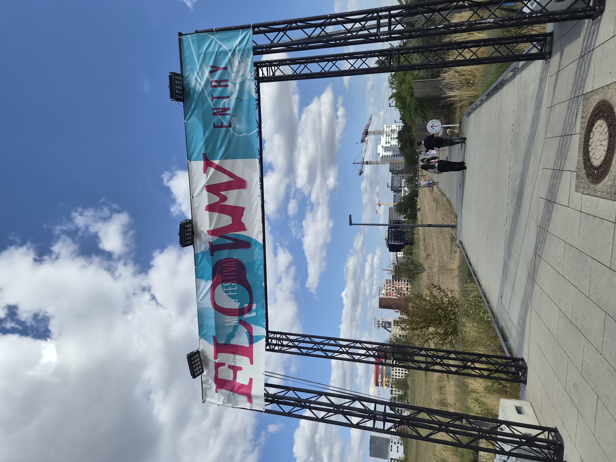 The image captures an outdoor scene beneath a partly cloudy blue sky. A large banner displaying "FLOW MUSIC FESTIVAL ENTRY" is suspended across a black metal archway. Two individuals are walking on a paved path leading away from the viewer, towards the banner. The path is bordered by dry grass and vegetation on the left and a metal fence on the right. In the background, a cityscape is visible with several buildings and construction cranes. The scene is located in the city of Sanem, Luxembourg.