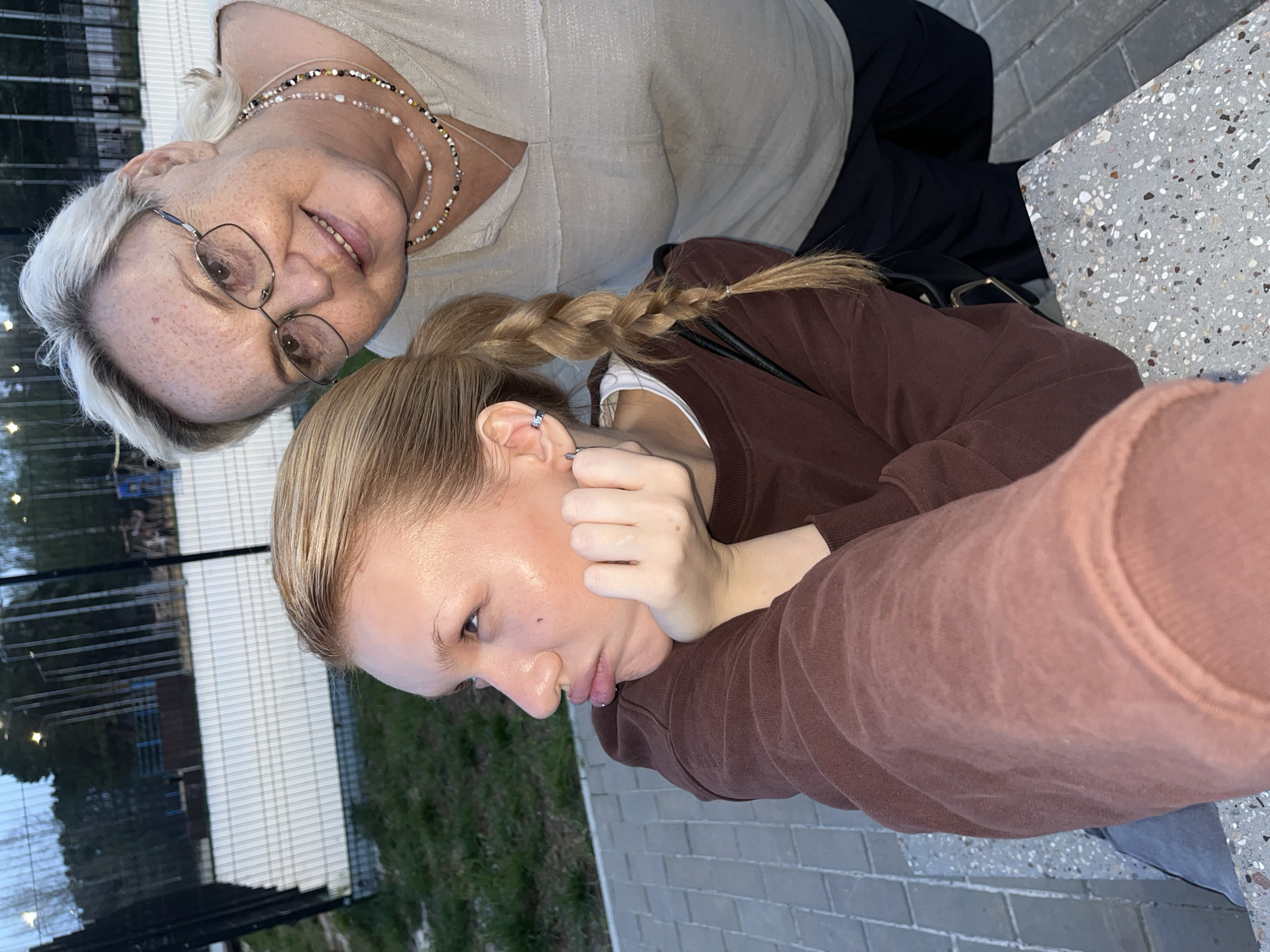 Two females pose in an outdoor setting in what appears to be Chaikovsky, Russia. One female is older with short white hair, wearing glasses, a light-colored top, and a multi-strand necklace. The other female is younger with blonde hair in a braid, wearing a brown sweatshirt. The younger female has a hand near her face. The surrounding environment includes brick paving, a metal fence, greenery, and a light-colored table surface.