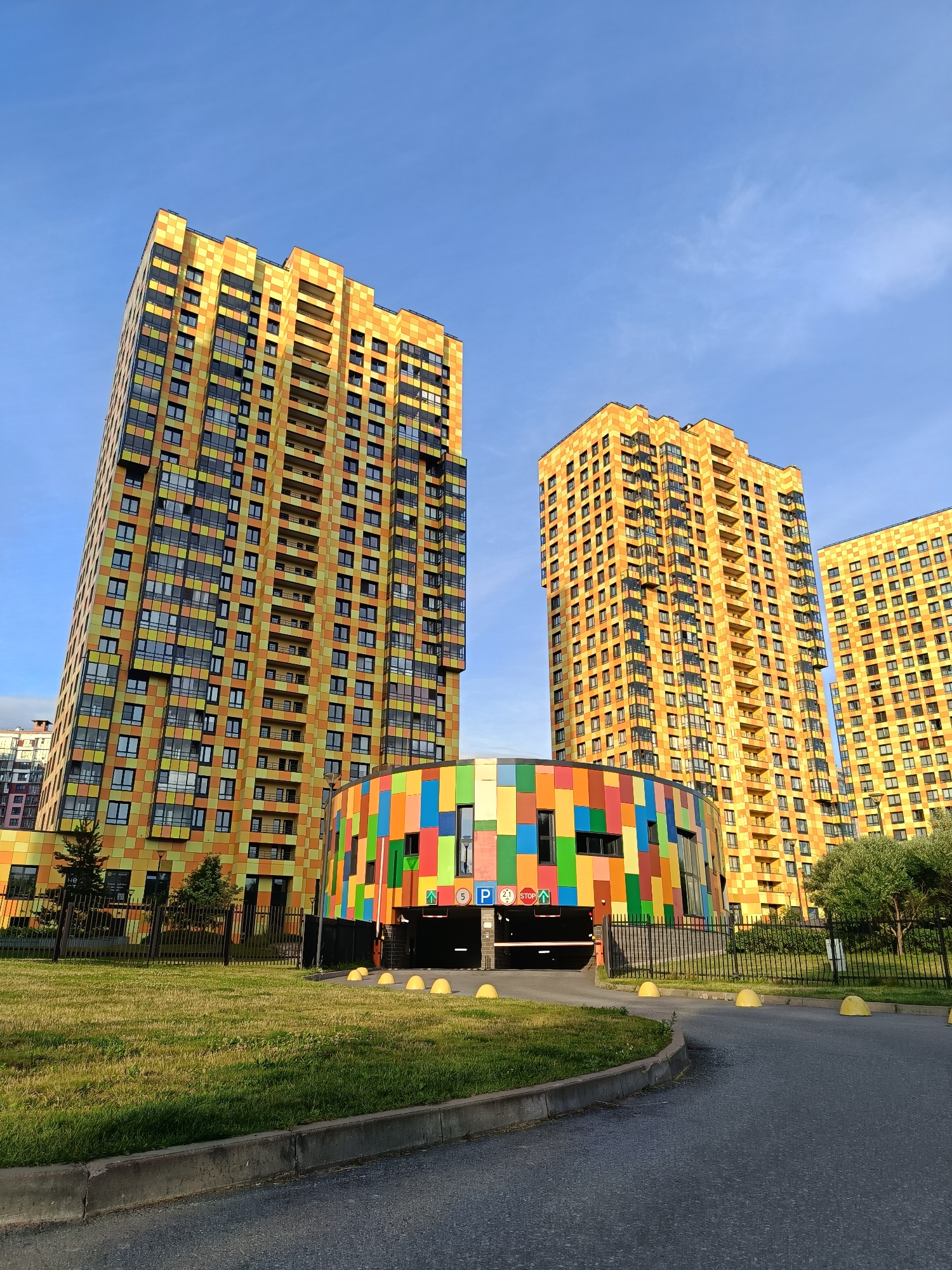 This is a low-angle outdoor, eye-level shot of a residential complex in Saint Petersburg, Russia. The architecture is modern and colorful, featuring two tall, orange and yellow residential buildings with a mosaic-like facade composed of square tiles in varying shades of yellow and orange. The buildings have numerous windows and balconies. In front of the buildings, a circular, multi-colored structure with a facade of colorful square panels in red, orange, yellow, green, blue, and purple serves as an entrance to an underground parking garage. The parking garage entrance is visible, with a barrier gate, and signage indicating "P" for parking, "STOP," and an upward arrow. Yellow hemispherical bollards are placed along the entrance to the parking garage. A black metal fence encloses the grassy area in front of the buildings. The ground is paved with asphalt, with a concrete curb. The sky above is clear blue with a few wispy clouds. The overall scene depicts a bright and vibrant urban residential environment.