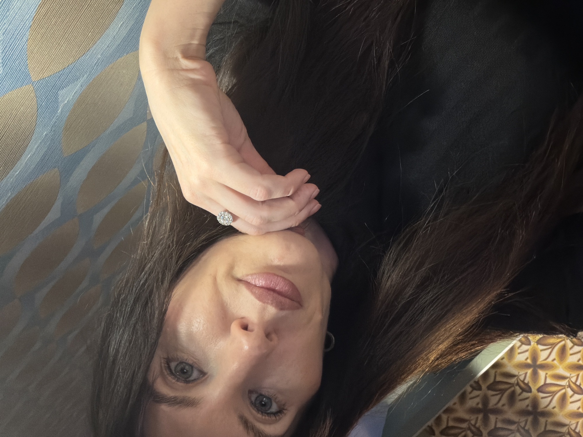 A woman with light skin, dark brown hair, and gray eyes is posing for a selfie. She is wearing a black top, and a ring with a large diamond is visible on her left hand. Her lips are lightly colored, and she has subtle makeup on her face. The background includes a blue and brown patterned surface on the left and a patterned surface on the lower right. The location is not specified in the image.